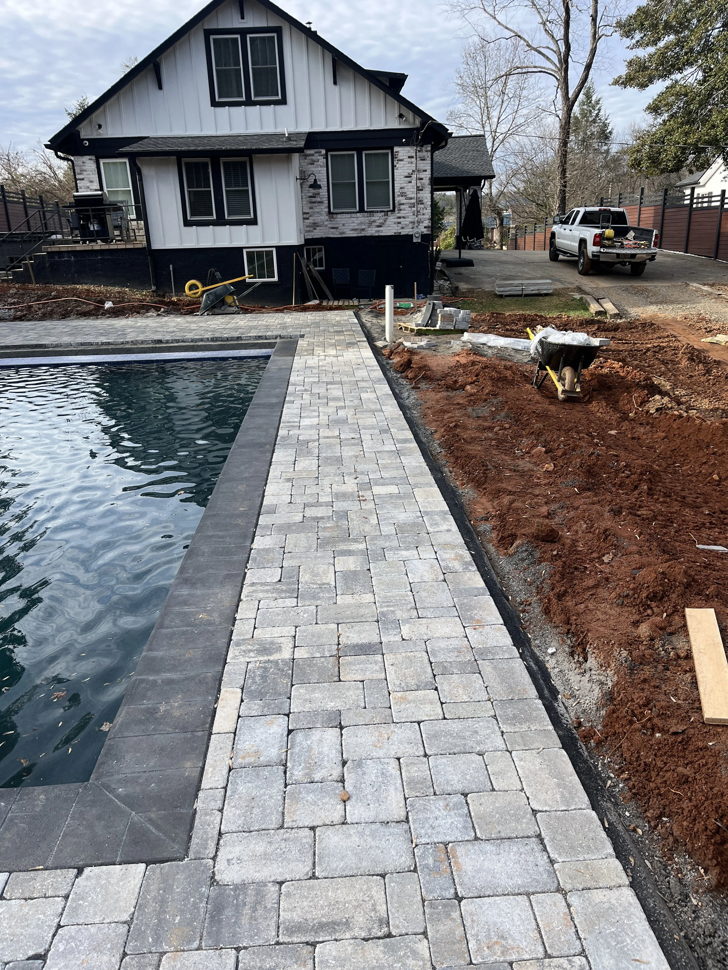 Construction site showing new paver walkway, a swimming pool, and a house in the background with ongoing landscaping work.