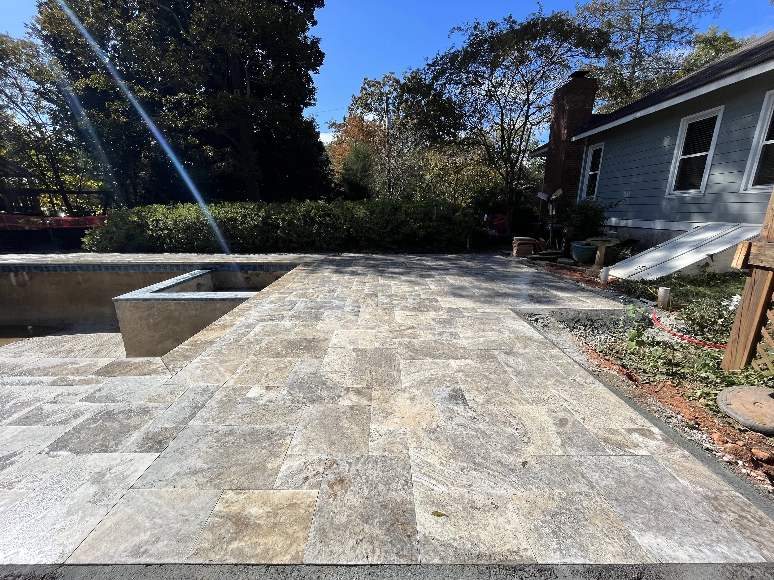 A backyard patio with a newly installed stone tile floor, a surrounding garden with bushes, trees, and a house with a chimney, windows, and a blue siding exterior. There is construction or landscaping work ongoing, as evidenced by the presence of a l