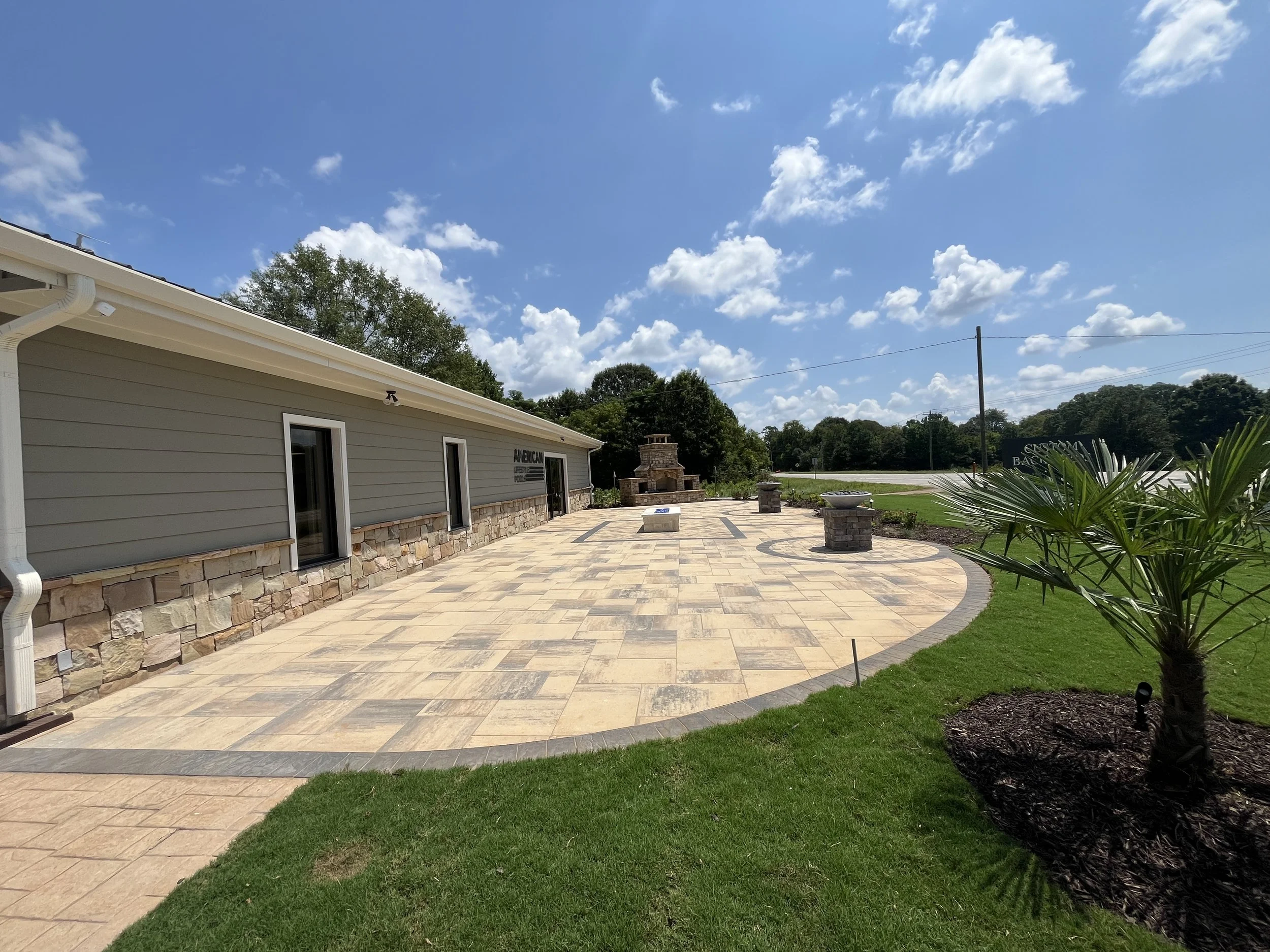 A backyard patio area with a stone and tile surface, adjacent to a house with light gray siding and stone accents. There are decorative planters, a brick fireplace, and well-maintained grass and plants. The sky is partly cloudy with a bright blue bac