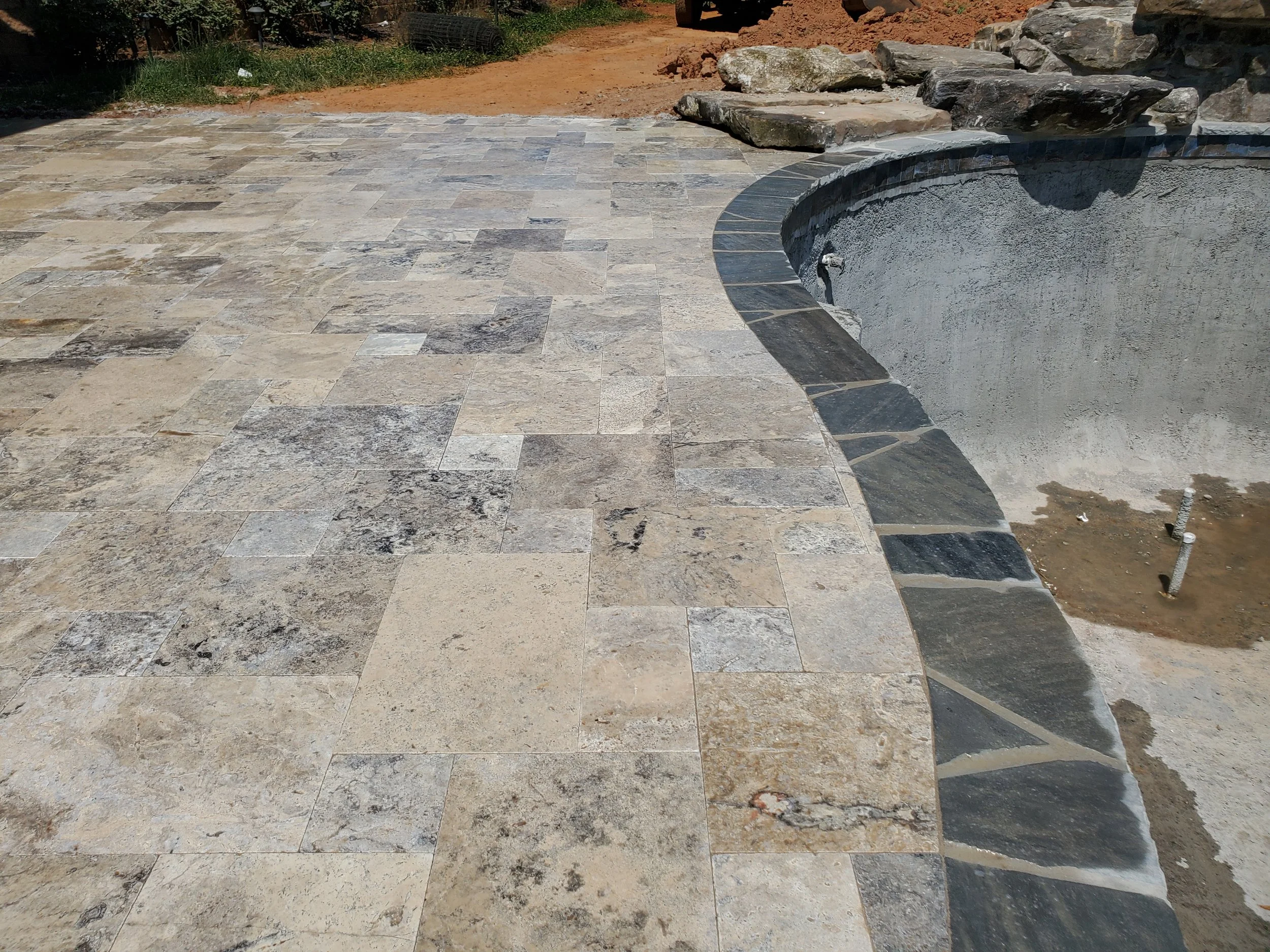 The image shows a section of a patio under construction, with newly laid stone tiles and a curved border. In the background, there is dirt, rocks, and some construction materials, indicating that work is ongoing.