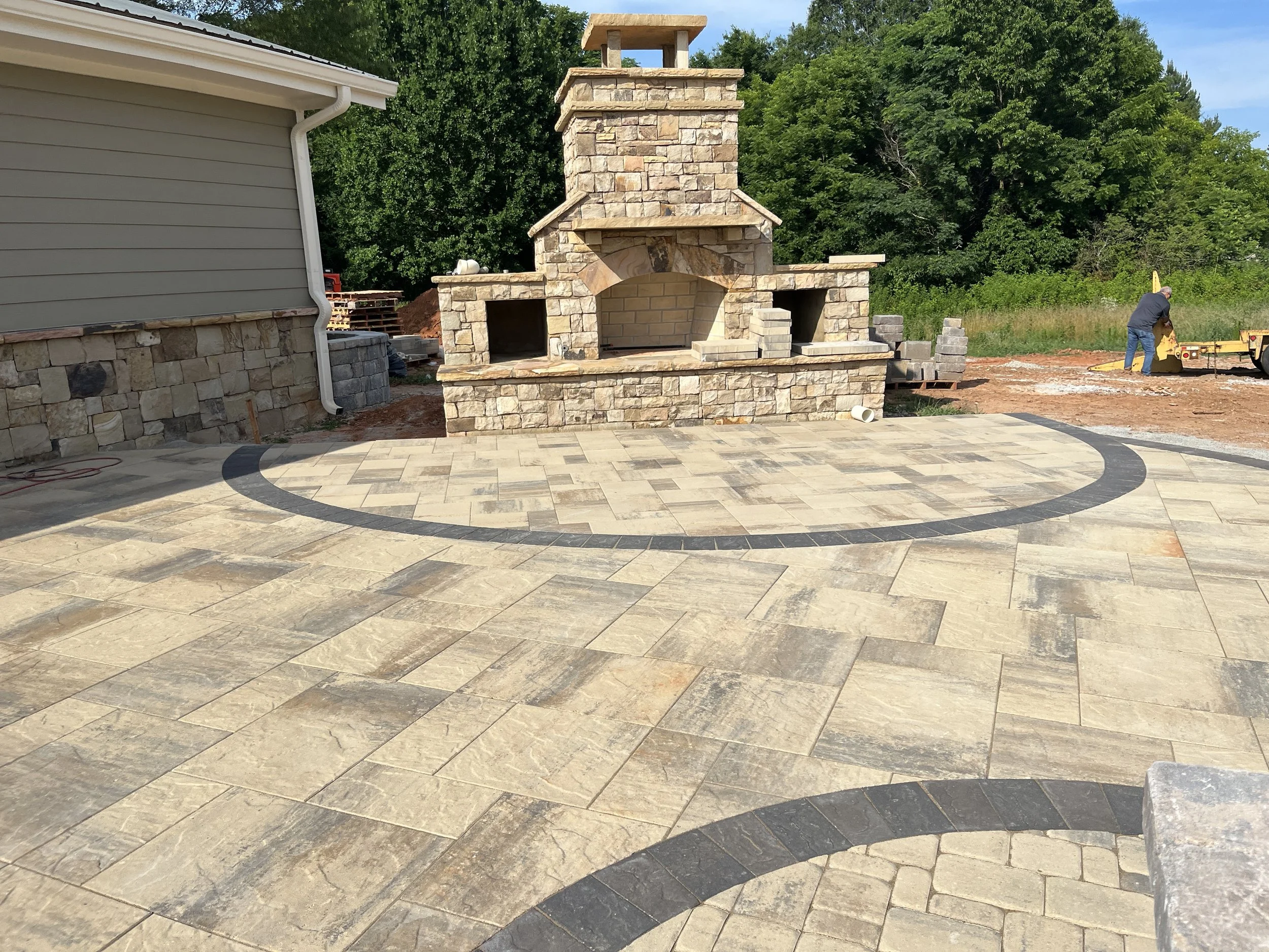 An outdoor patio area with a stone fireplace and a paved surface. A construction worker is working on a yellow piece of equipment in the background, with trees and a blue sky behind them.