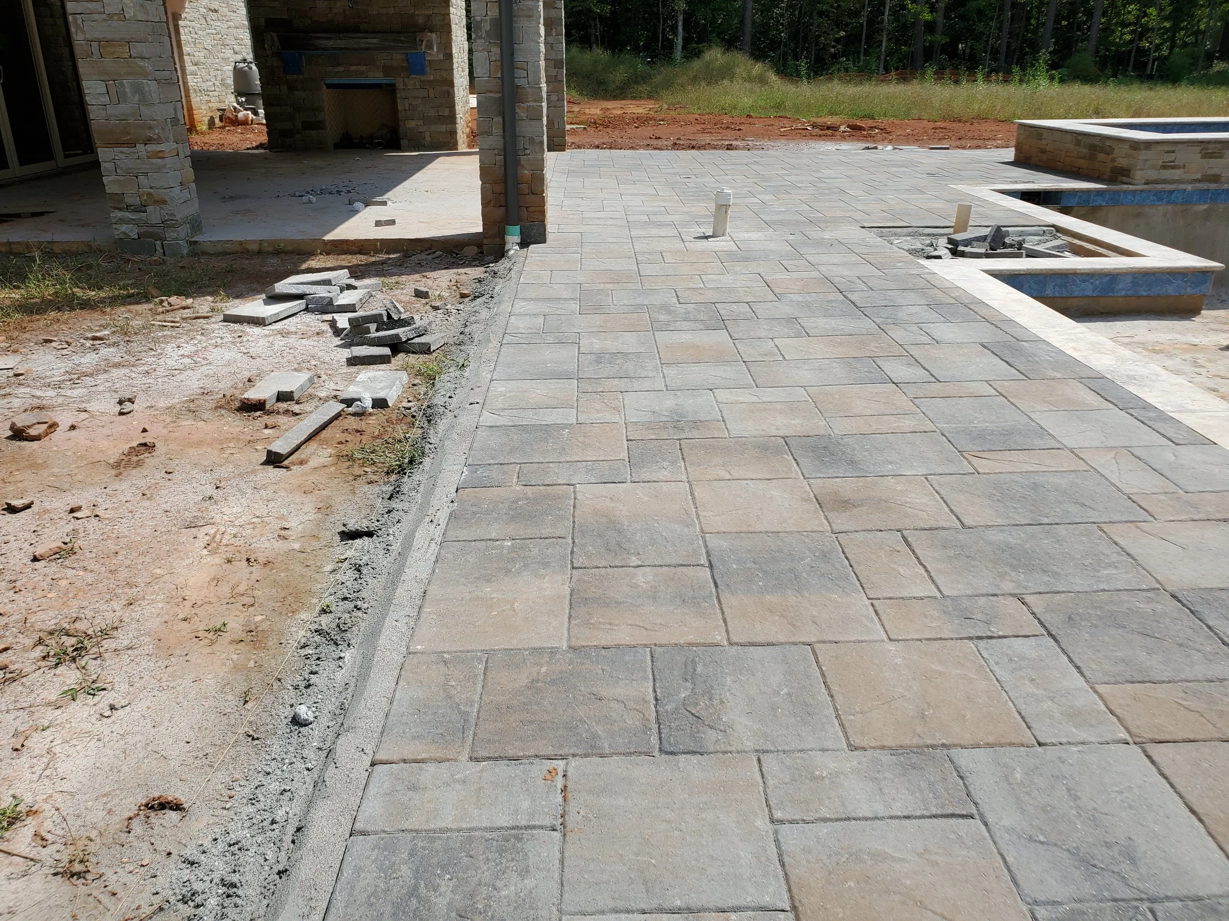 A backyard patio area under construction with a paved stone surface, a raised pool with blue tiles, a brick fireplace, and a partially finished area with construction debris and tools.