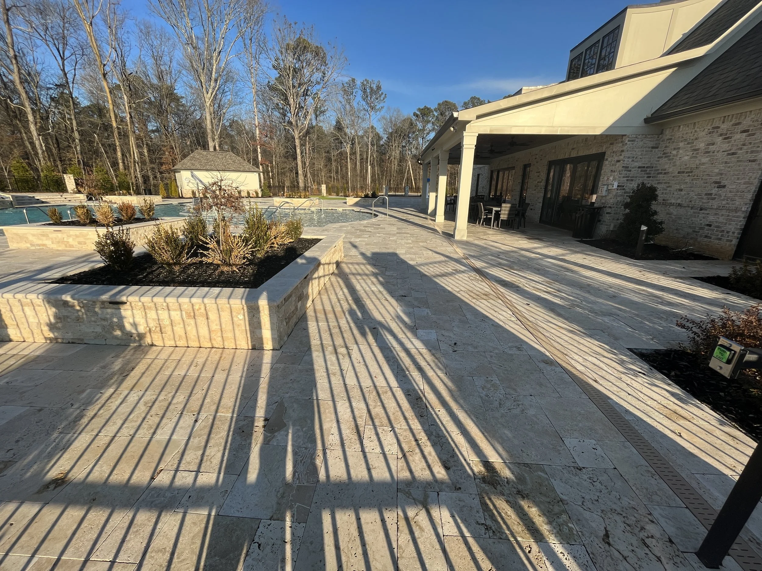 Spacious outdoor patio area with stone paving, a swimming pool, outdoor seating, and a view of leafless trees under a clear blue sky.