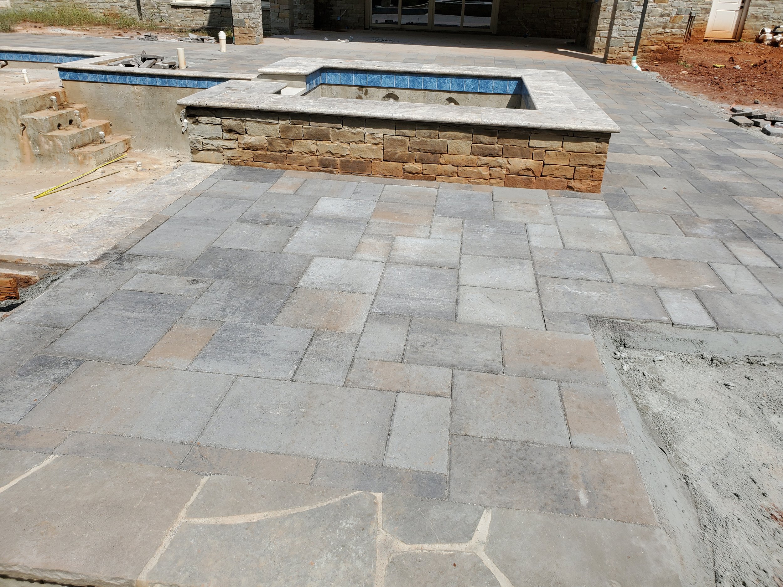 Construction site showing a partially built in-ground pool with blue tile accents, surrounded by gray stone paving and some construction debris.