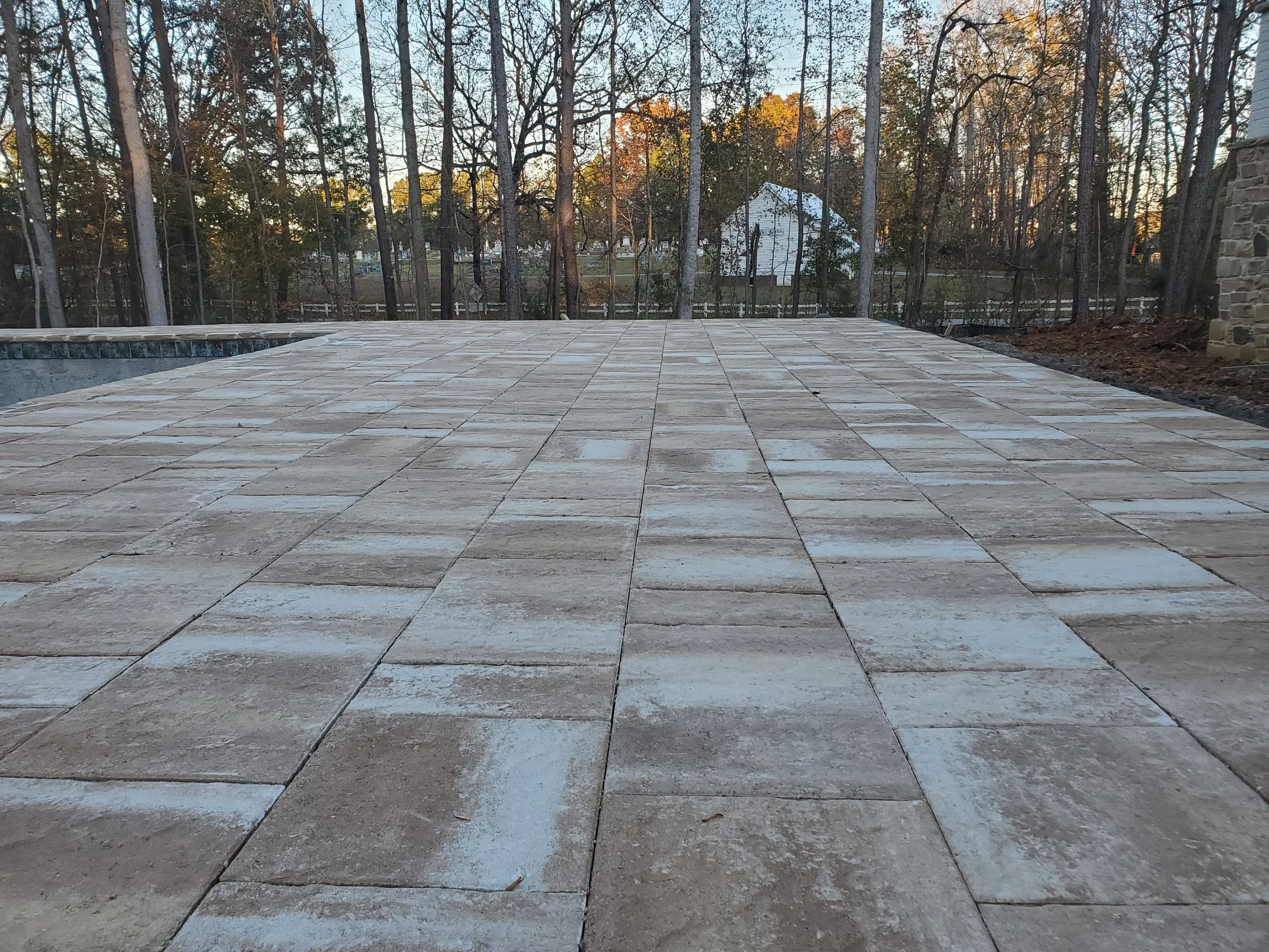 A newly built stone patio surrounded by a wooded area with trees and a white barn or shed in the distance, under a clear sky at sunset.