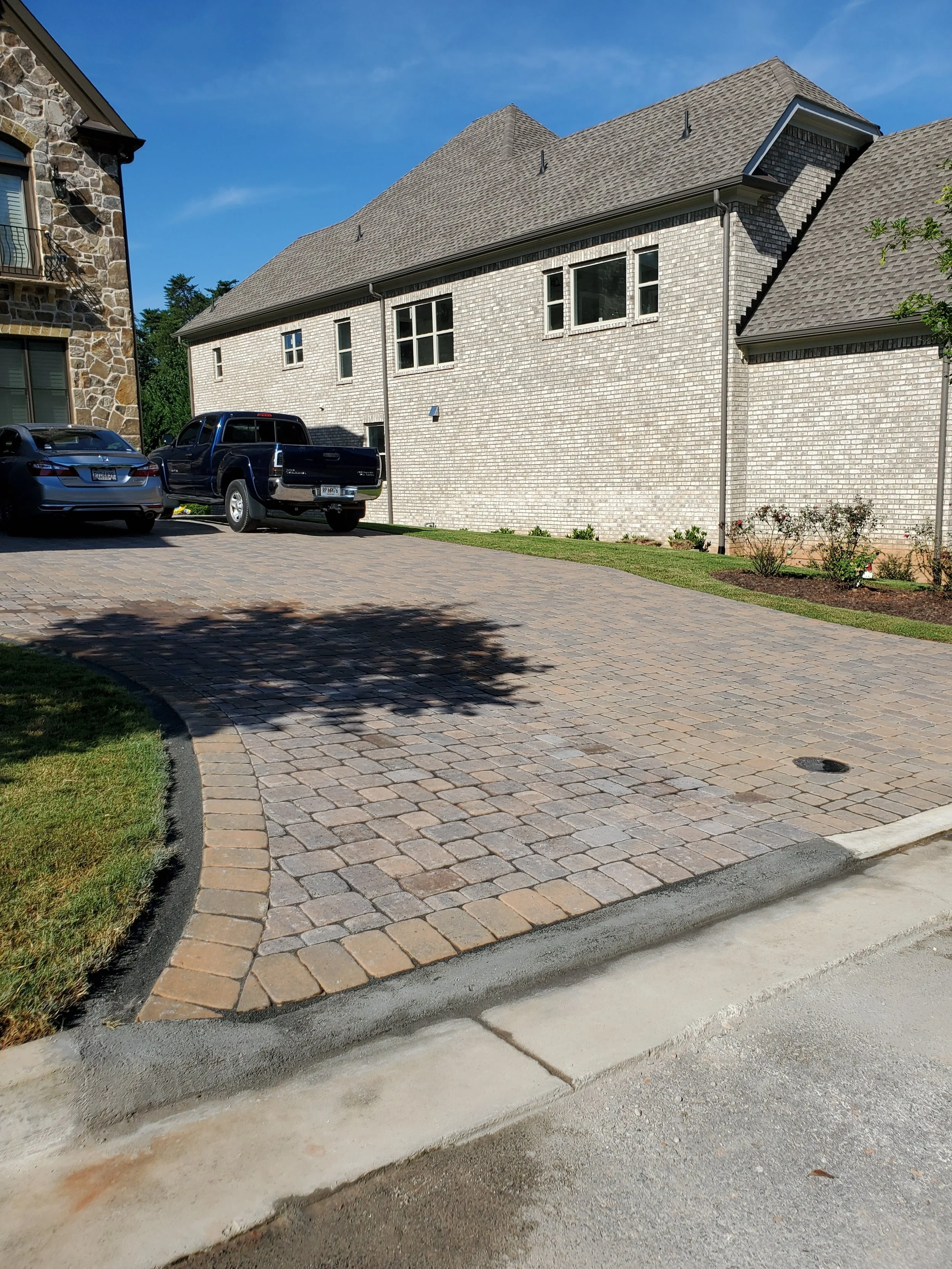 A newly paved brick driveway curves away from a house with a white brick exterior and a gray roof, with two cars parked on it and a small garden bed with flowers along the side.