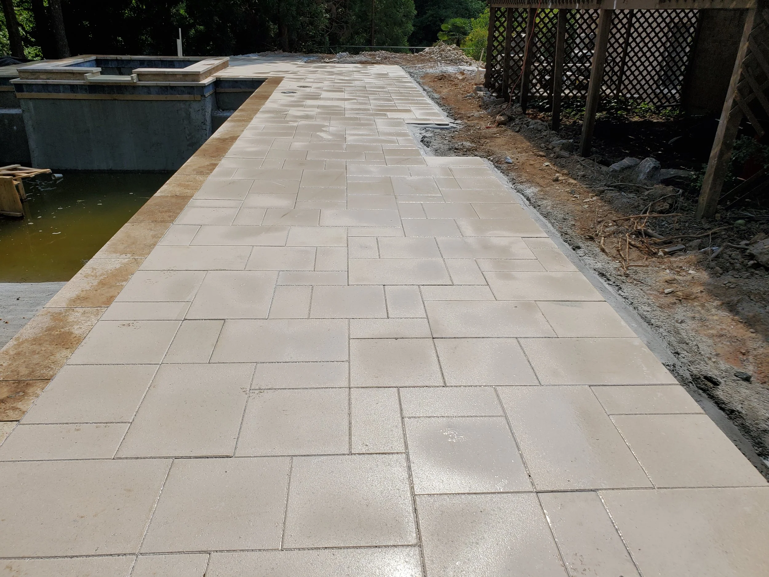 Newly paved light-colored stone patio with a small raised hot tub at the left end, adjacent to a pond or water feature, in a backyard setting with surrounding trees and fencing.
