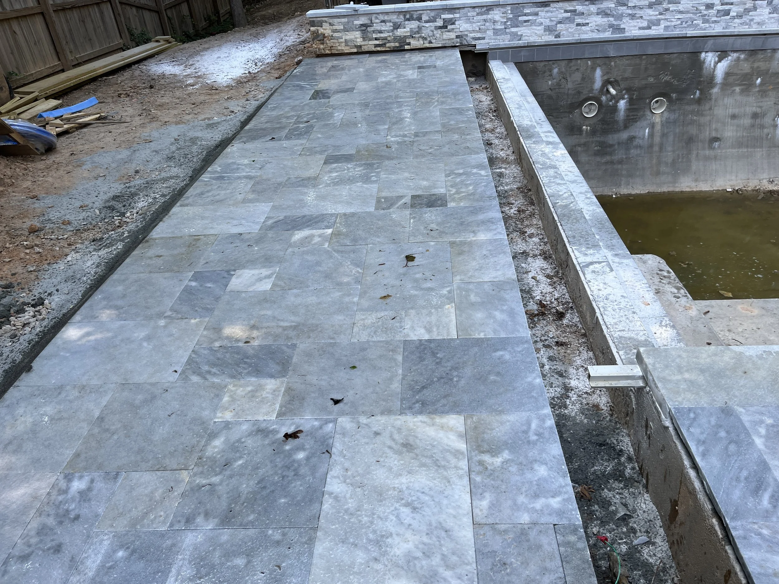 Construction site with new gray stone pavers installed on walkway, adjacent to a pool with unfinished interior and pipes visible, surrounded by dirt, wood piles, and construction materials.