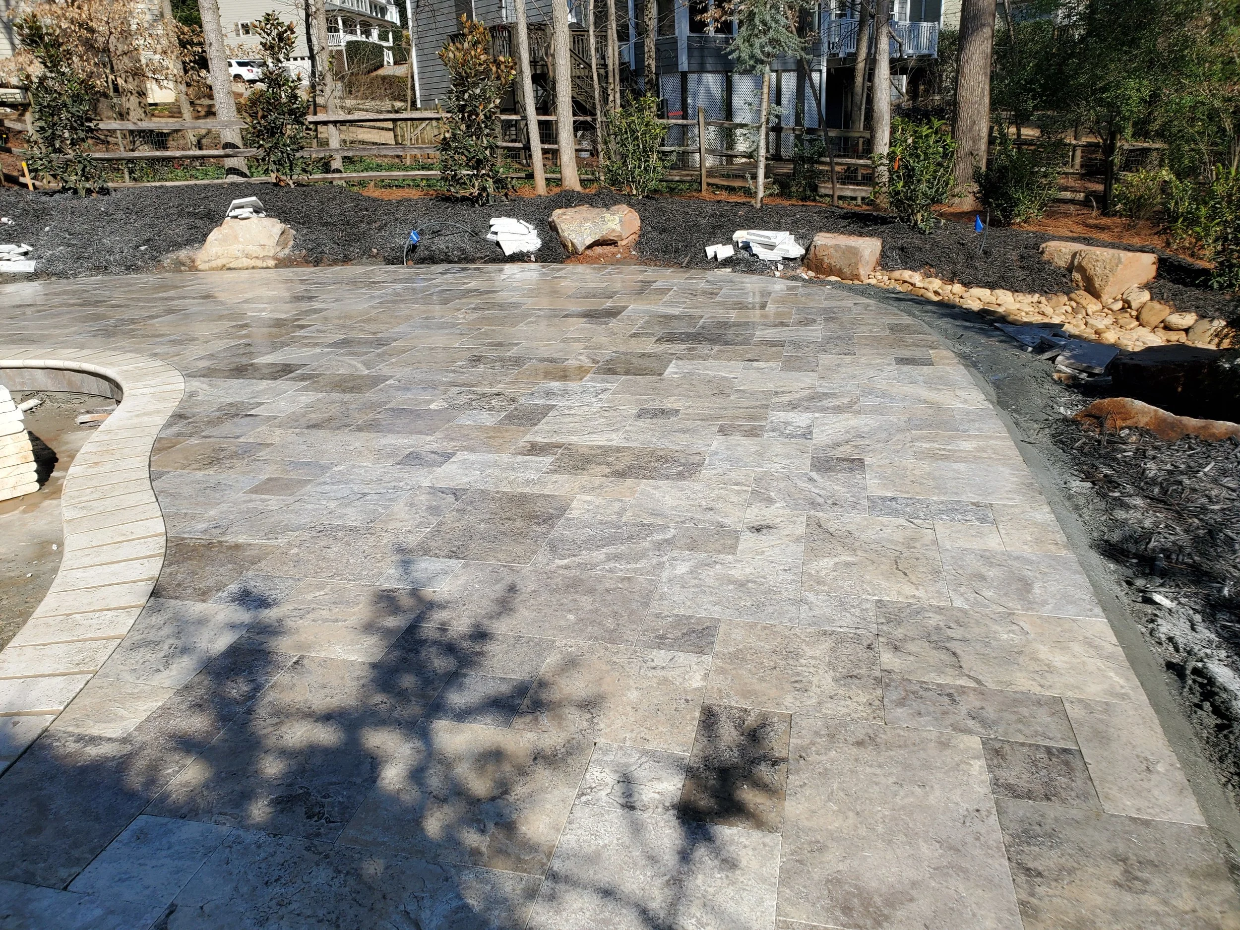 A backyard patio with a stone tile surface, several large rocks, and small flag markers in a garden area with trees, bushes, and a wooden fence.