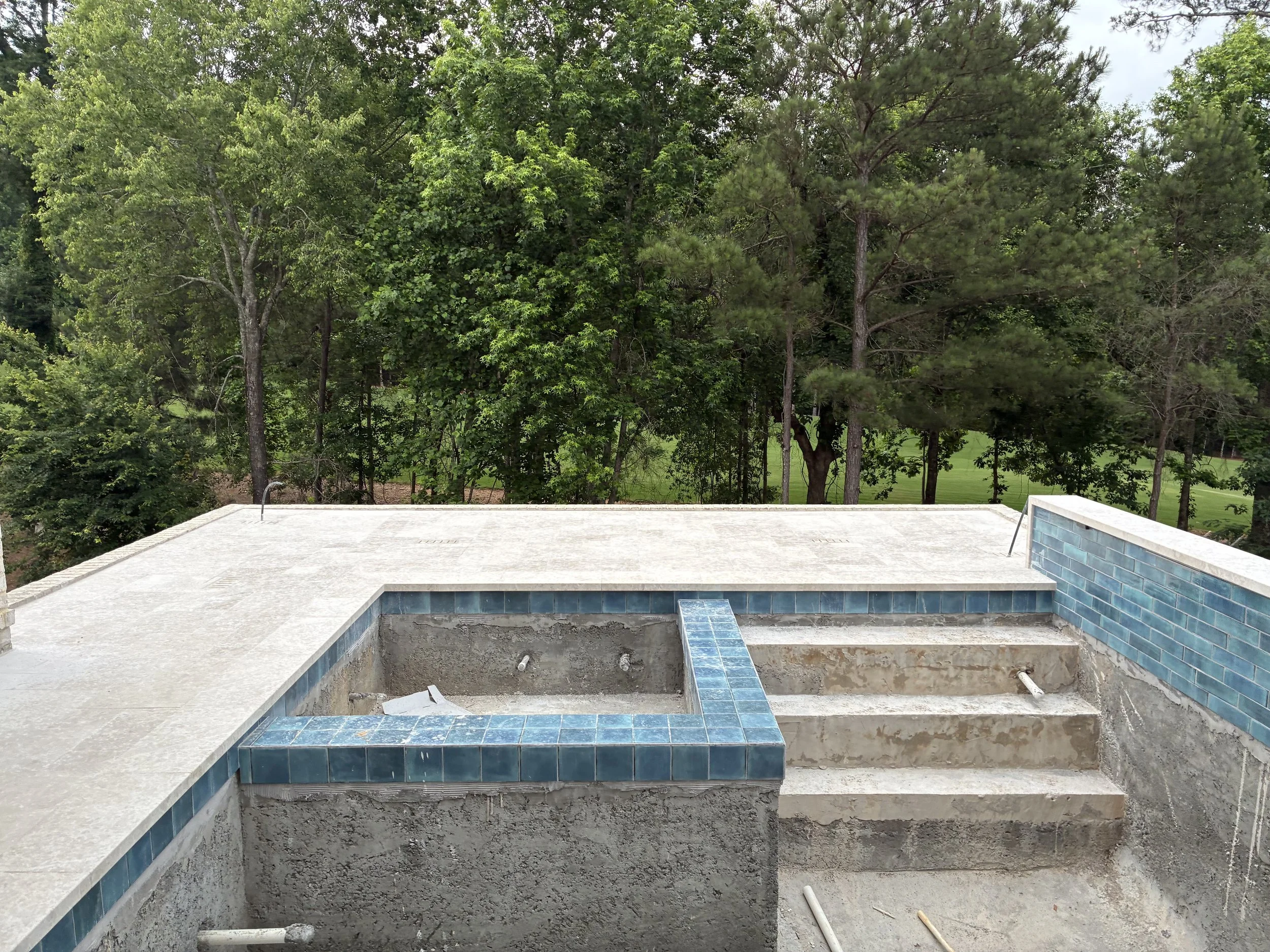 An outdoor swimming pool under construction with blue tile lining and a concrete deck, surrounded by green trees.