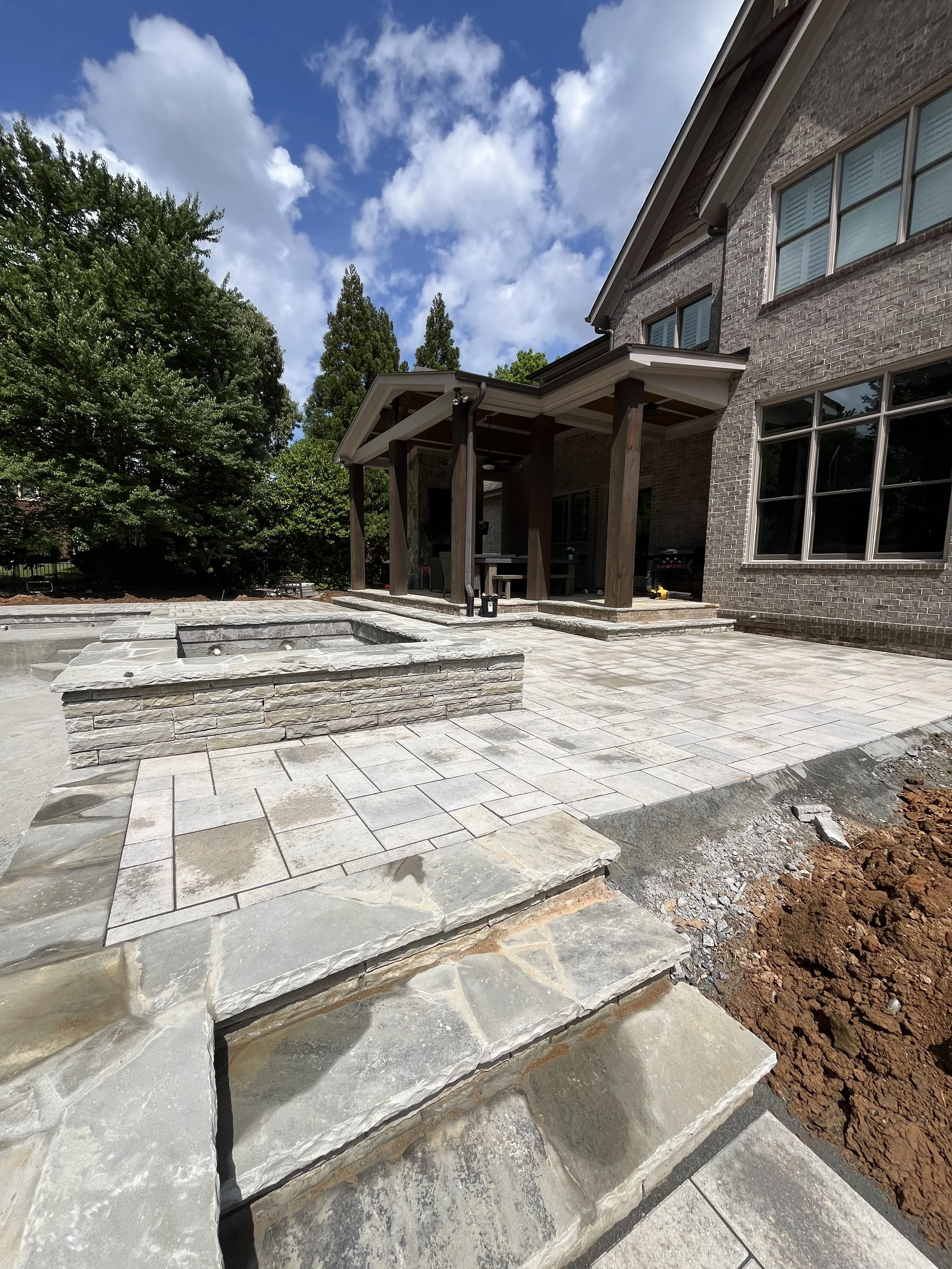 An outdoor patio with stone steps and paving, part of a brick house, under a partly cloudy sky with trees in the background.