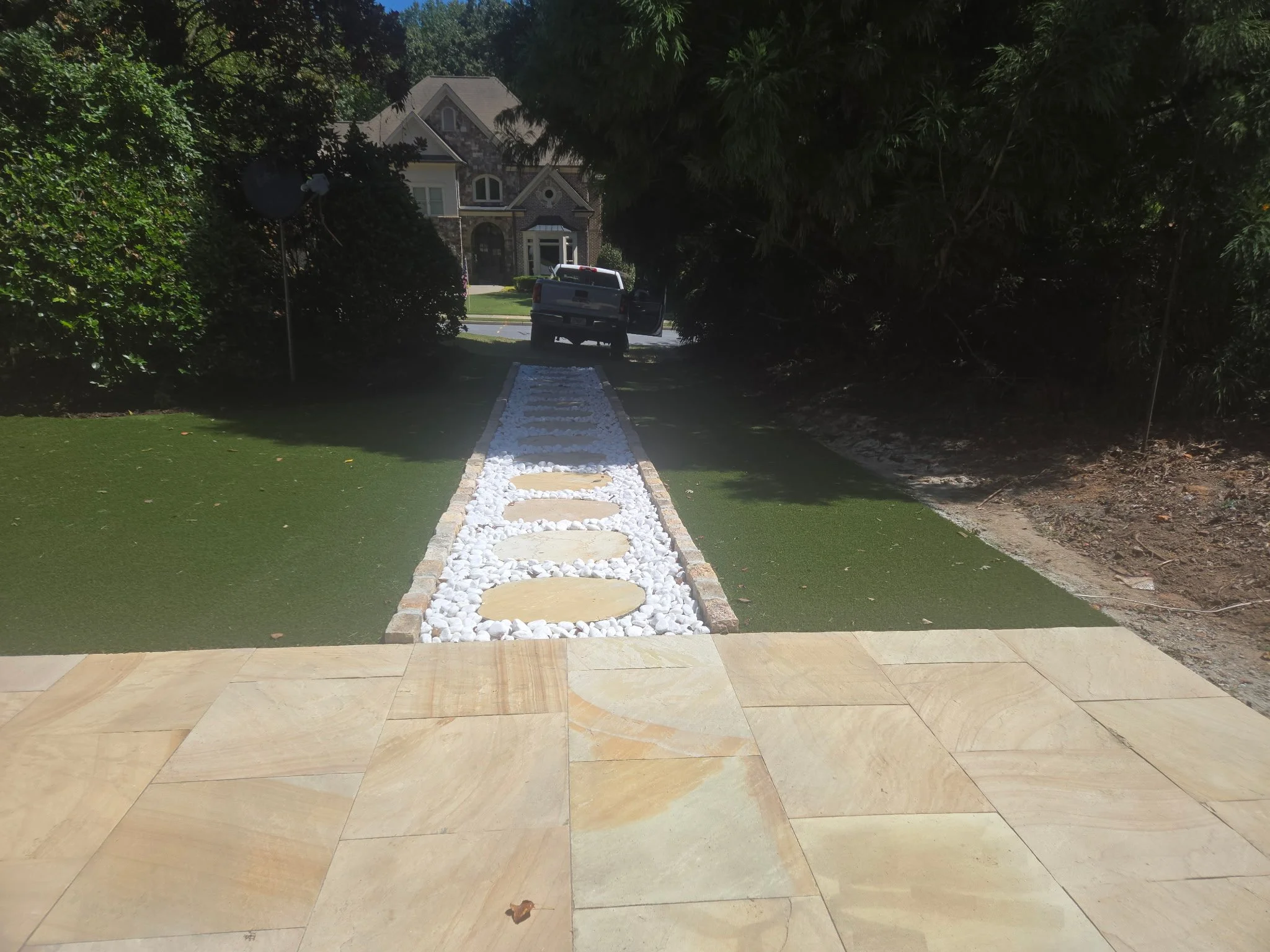 A landscaped front yard with a stone pathway leading to a house, flanked by green grass and bushes, with a truck parked in front of the house.