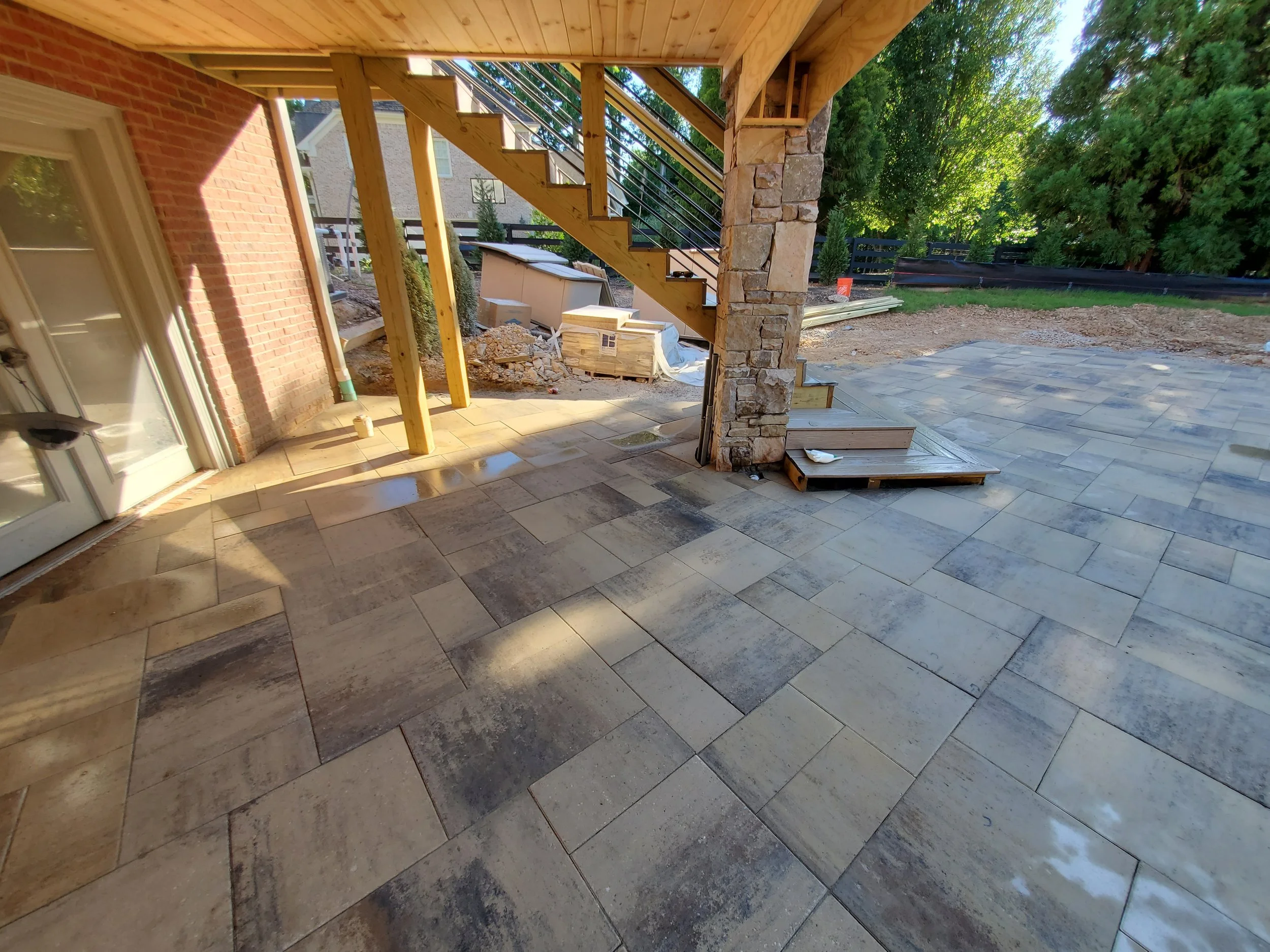 Under construction outdoor patio area with stone and tile flooring, wooden staircase, and partial brick wall, surrounded by greenery.