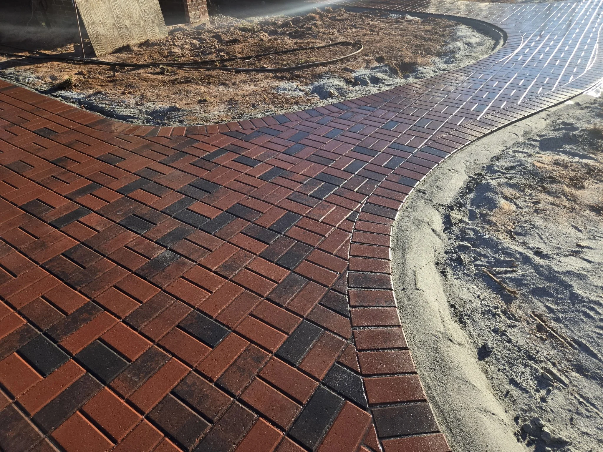 Red brick sidewalk with curving pattern, partially finished with sand and dirt adjacent to it, and construction tools visible, indicating ongoing paving work.