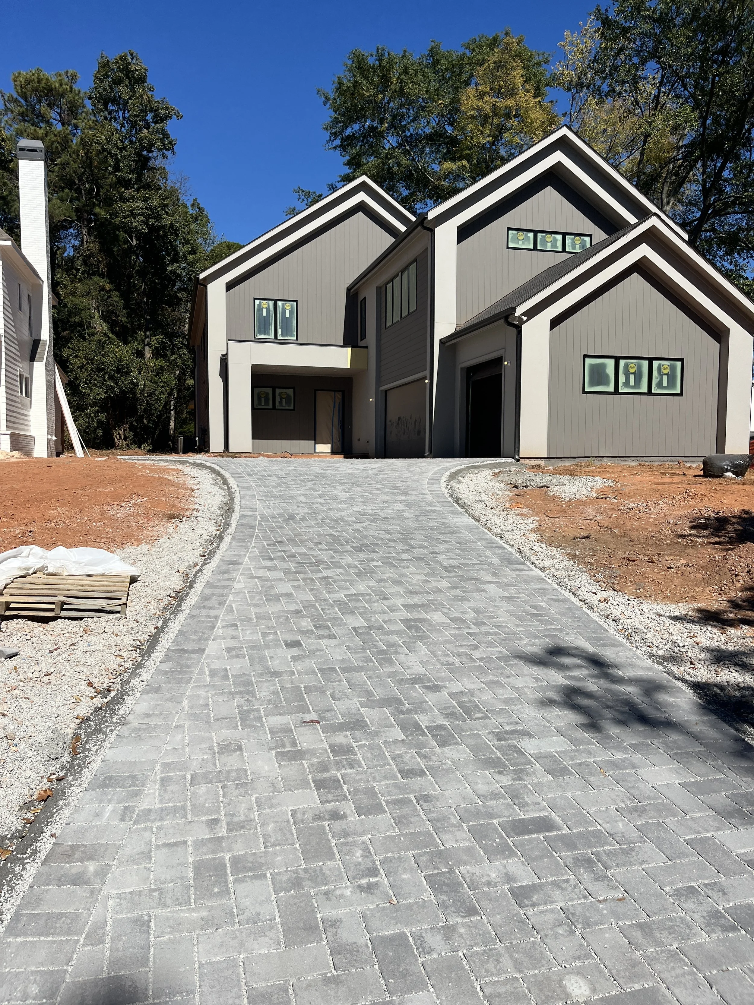Newly paved brick driveway leading to modern house with grey siding and black trim, surrounded by trees.