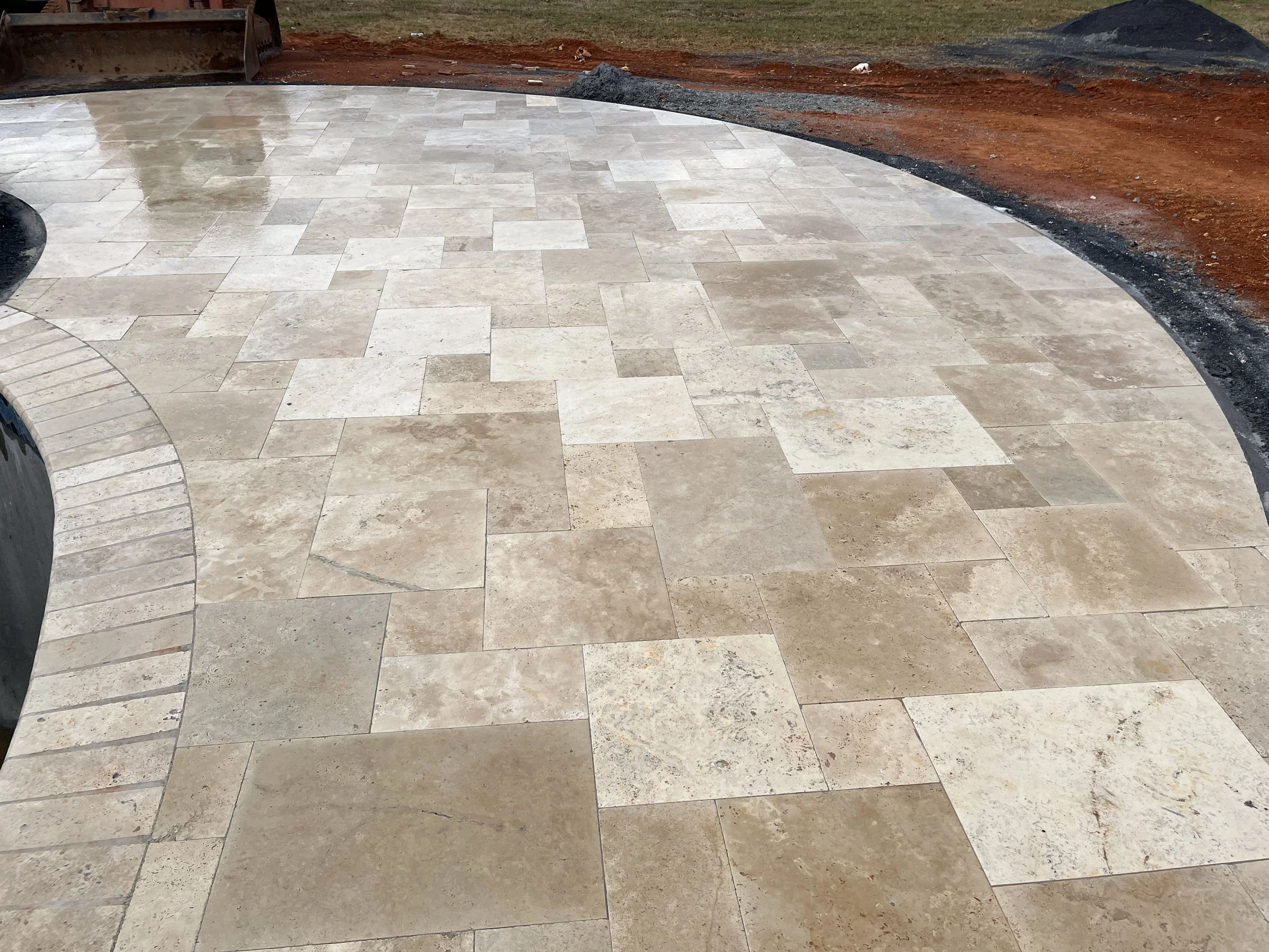 Newly installed beige and light brown stone patio with a curved edge, surrounded by prepared red soil and construction materials, indicating ongoing outdoor landscaping work.