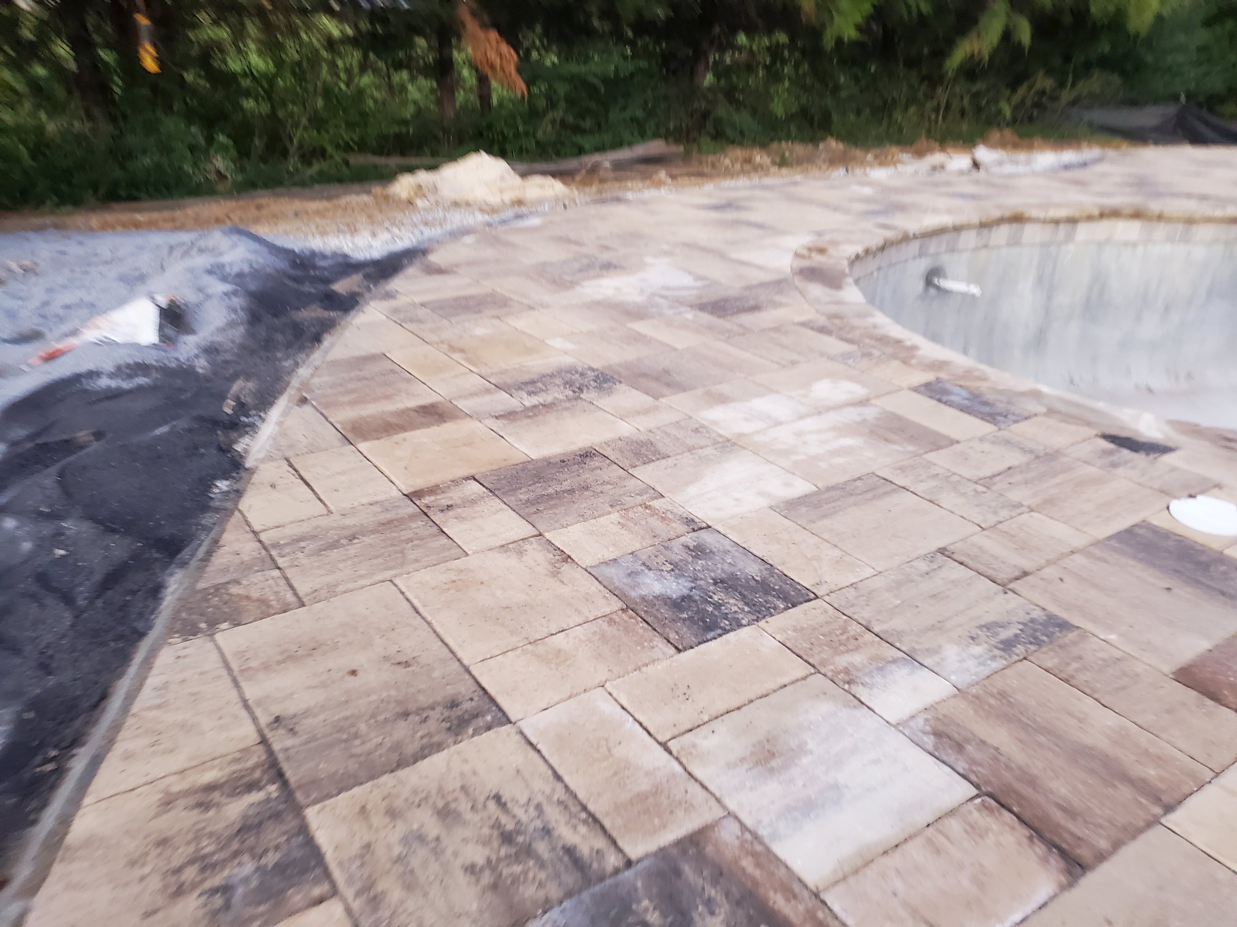 Partially completed brick paved patio with a curved edge, some tools and materials scattered around, and a background of green shrubbery and trees.