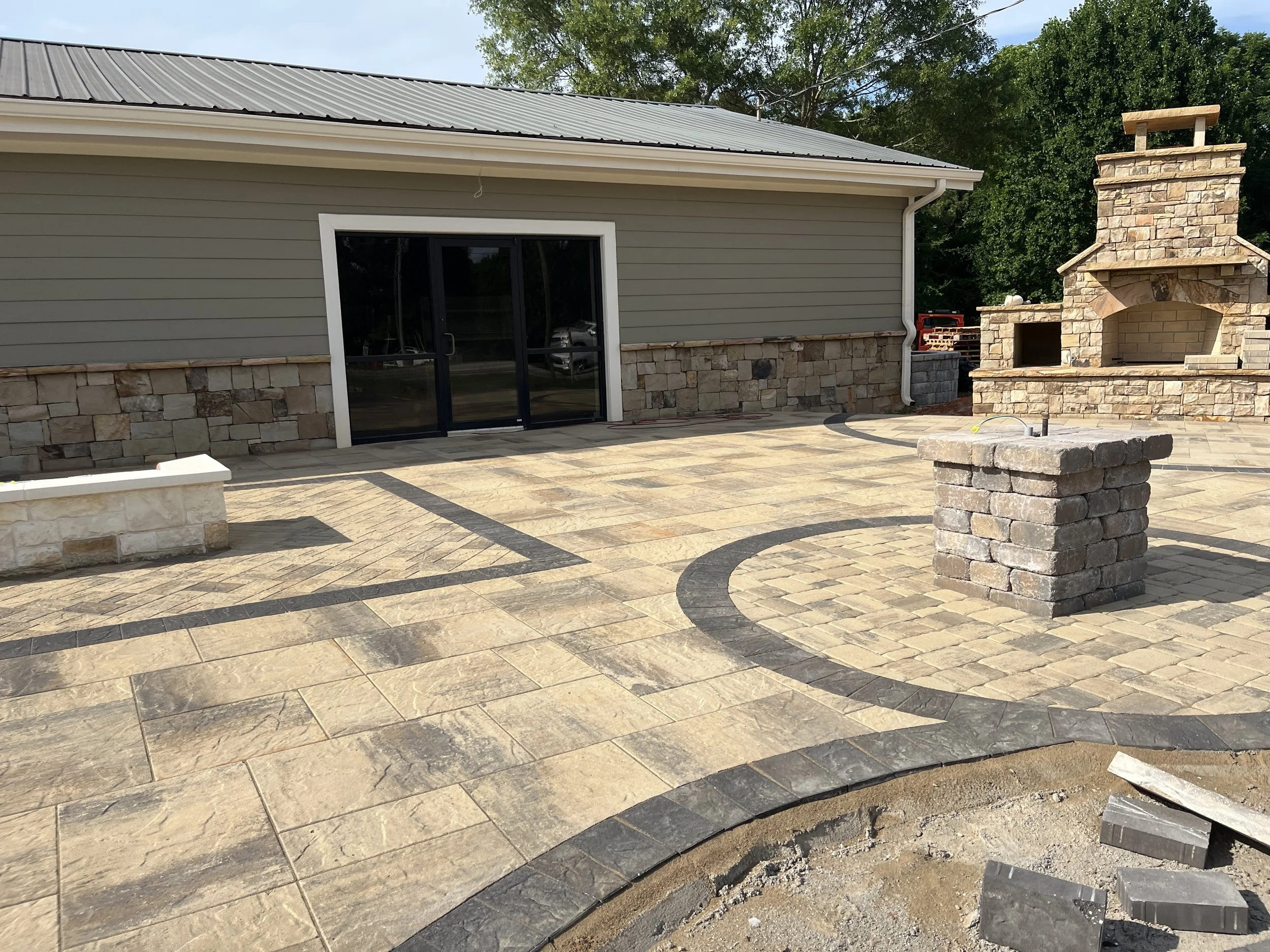 Backyard patio area with light-colored stone tiles, a stone outdoor fireplace, and a sliding glass door leading into the house. There are trees and greenery in the background.