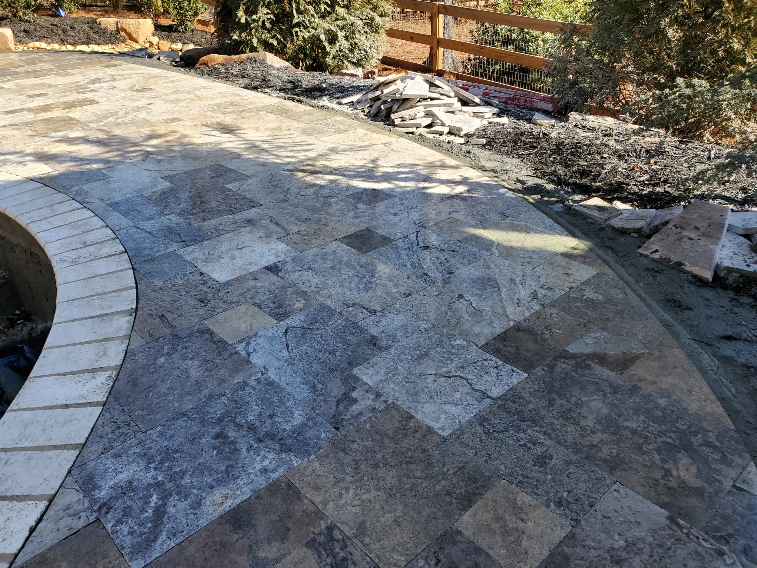 A section of a stone patio with a curved border, next to a landscaped area with mulch, rocks, and bushes. In the background, there is a wood fence and trees.