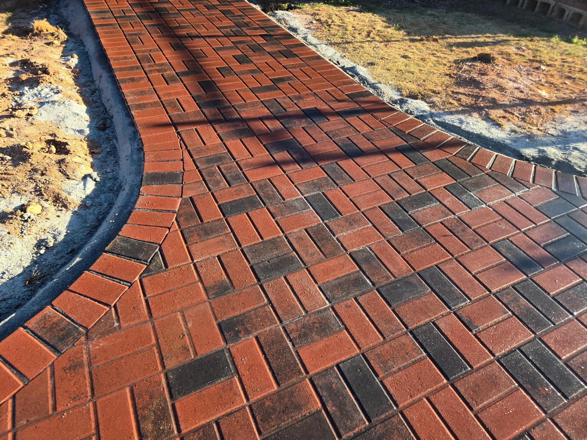 A freshly laid brick sidewalk with a curved edge, bordered by dirt and grass, taken during daylight.