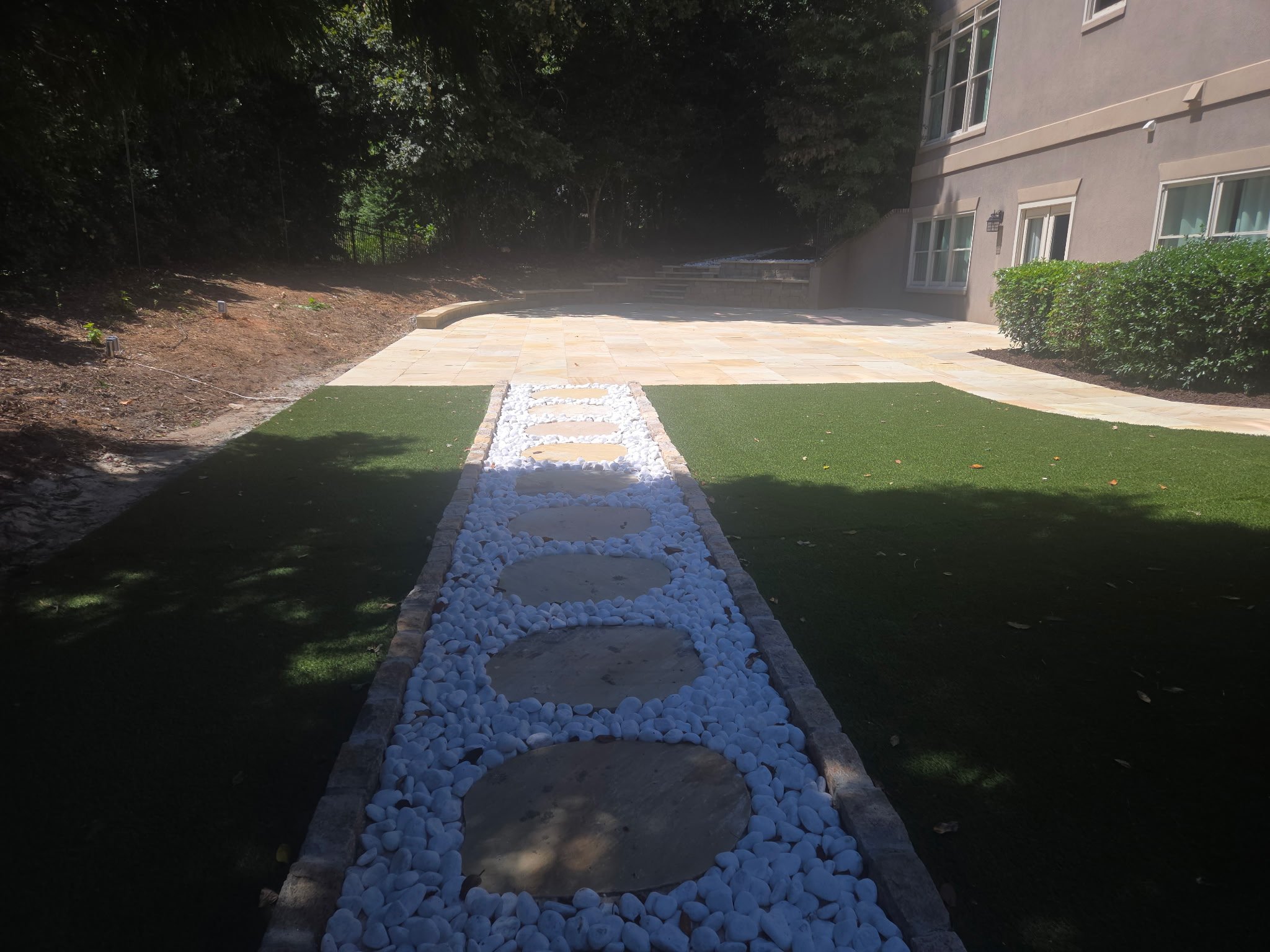 A backyard patio with a stepping stone pathway bordered by white rocks, adjacent to a synthetic lawn.