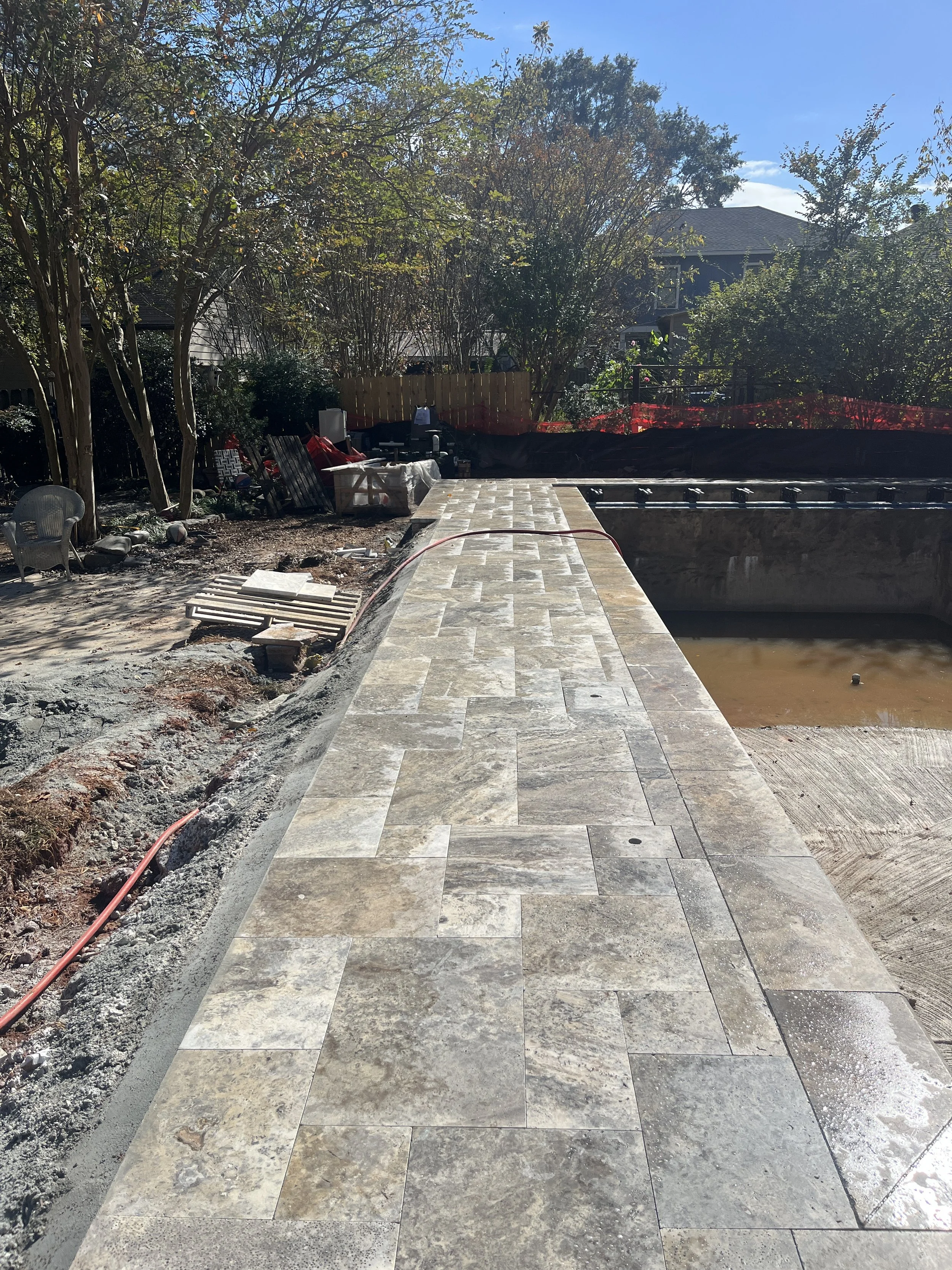 Backyard with a partially completed stone patio, construction materials, and outdoor furniture, with trees and a house in the background under a clear blue sky.