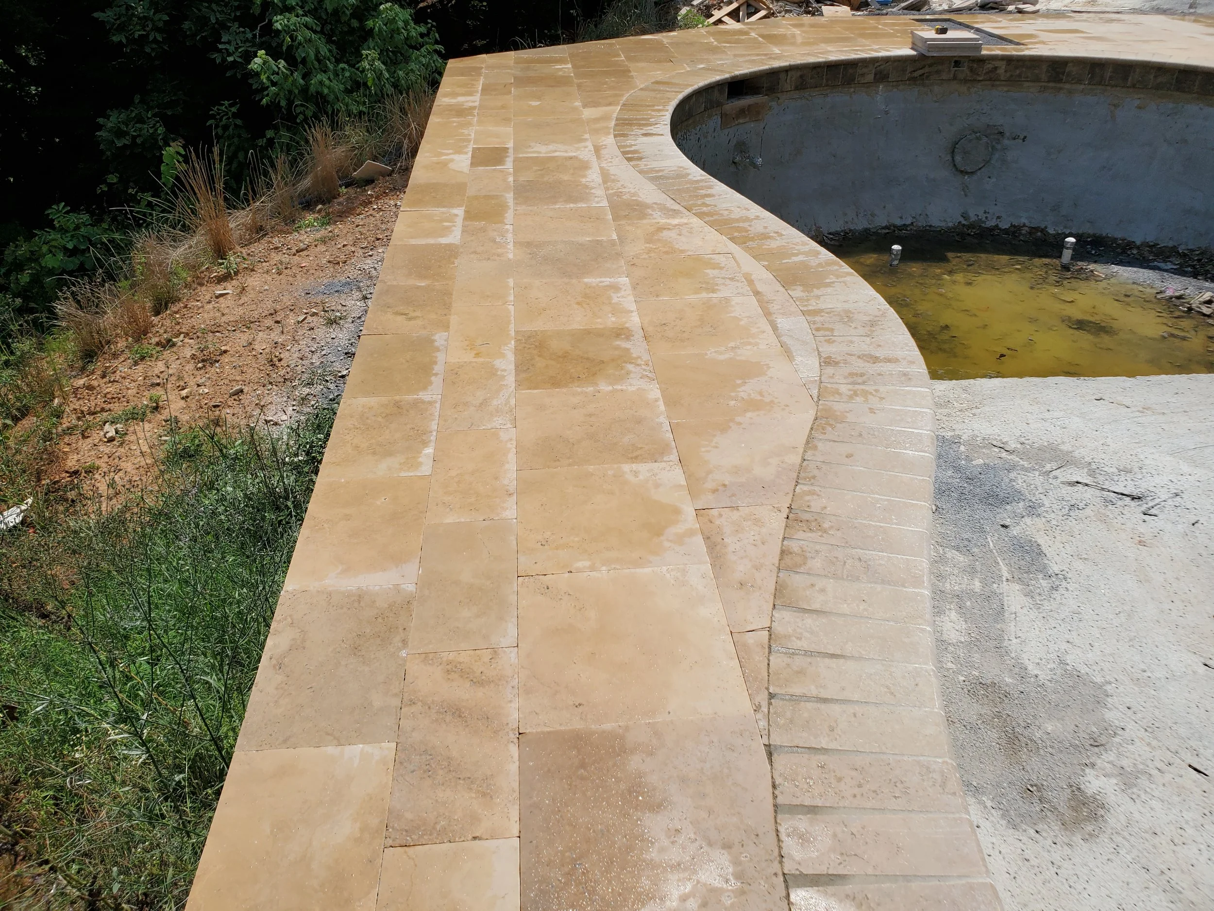 A backyard with a partially completed swimming pool. The pool has a curved shape with beige tile coping and a gray interior. The surrounding area is muddy with sparse grass and weeds, and there is green foliage in the background.