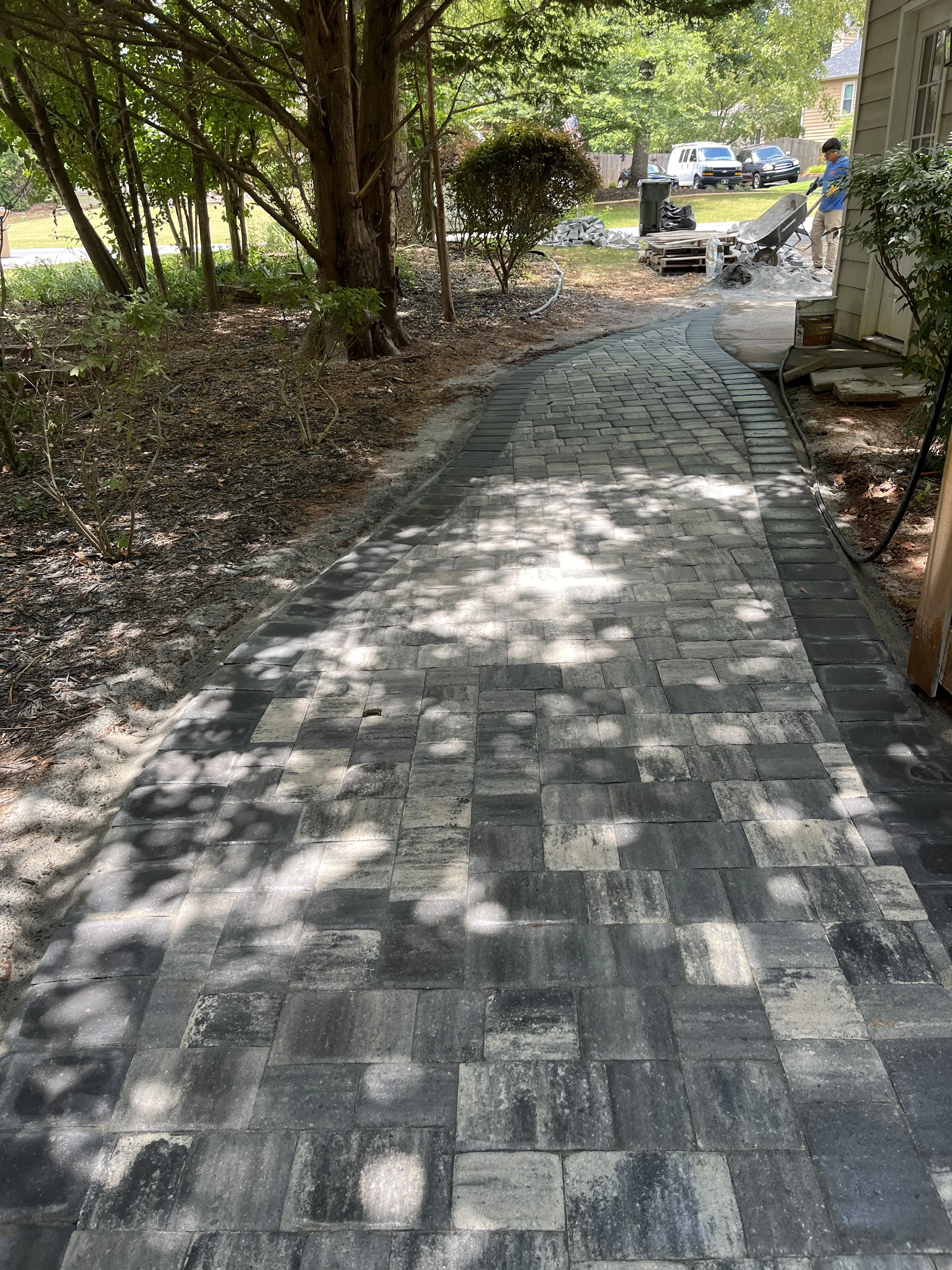 A person working on laying down a new brick pathway in a backyard, shaded by trees, with house and cars visible in the background.