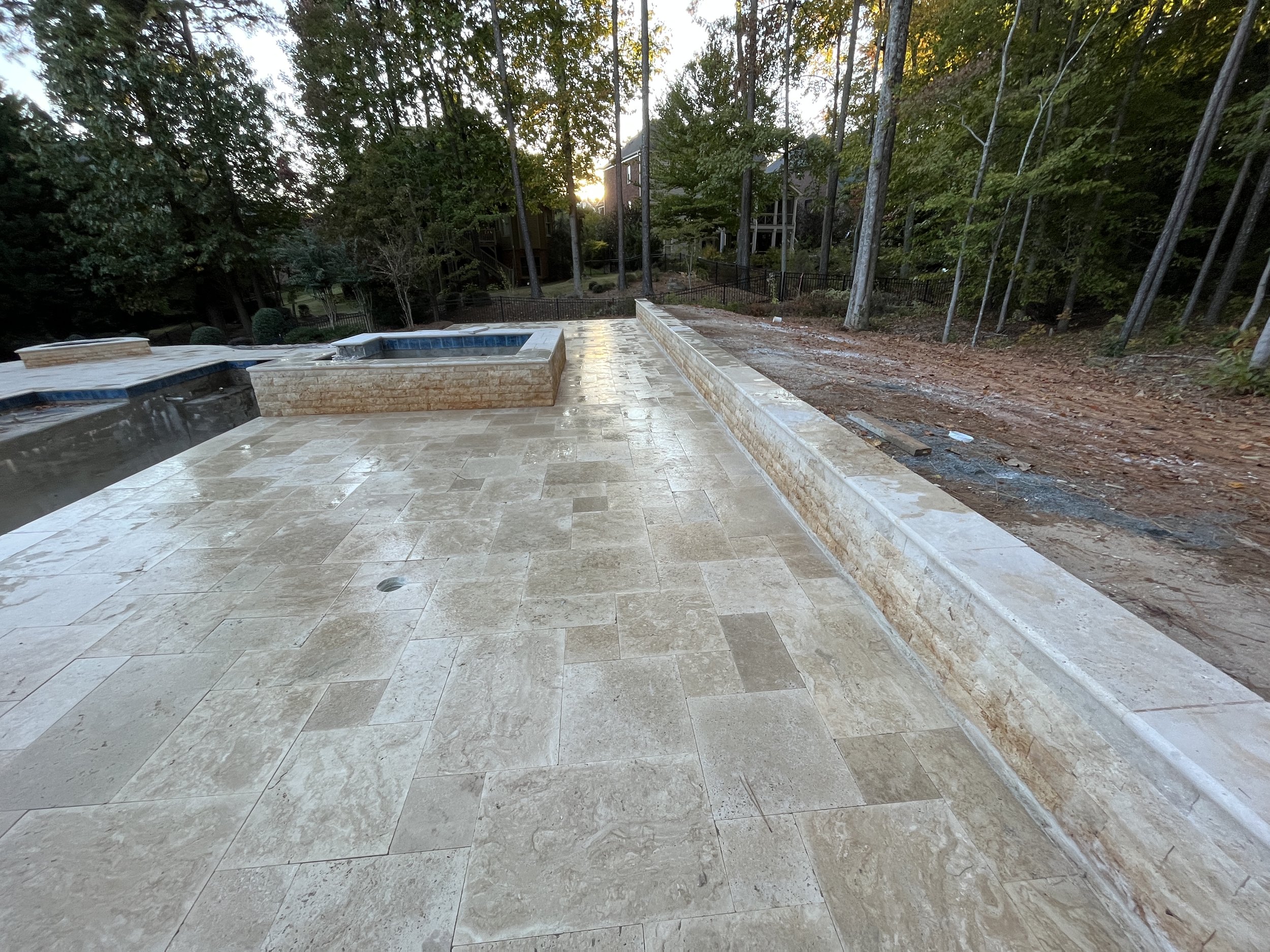 A backyard with a stone patio, a built-in hot tub, and surrounded by trees with houses in the background and the sun setting.