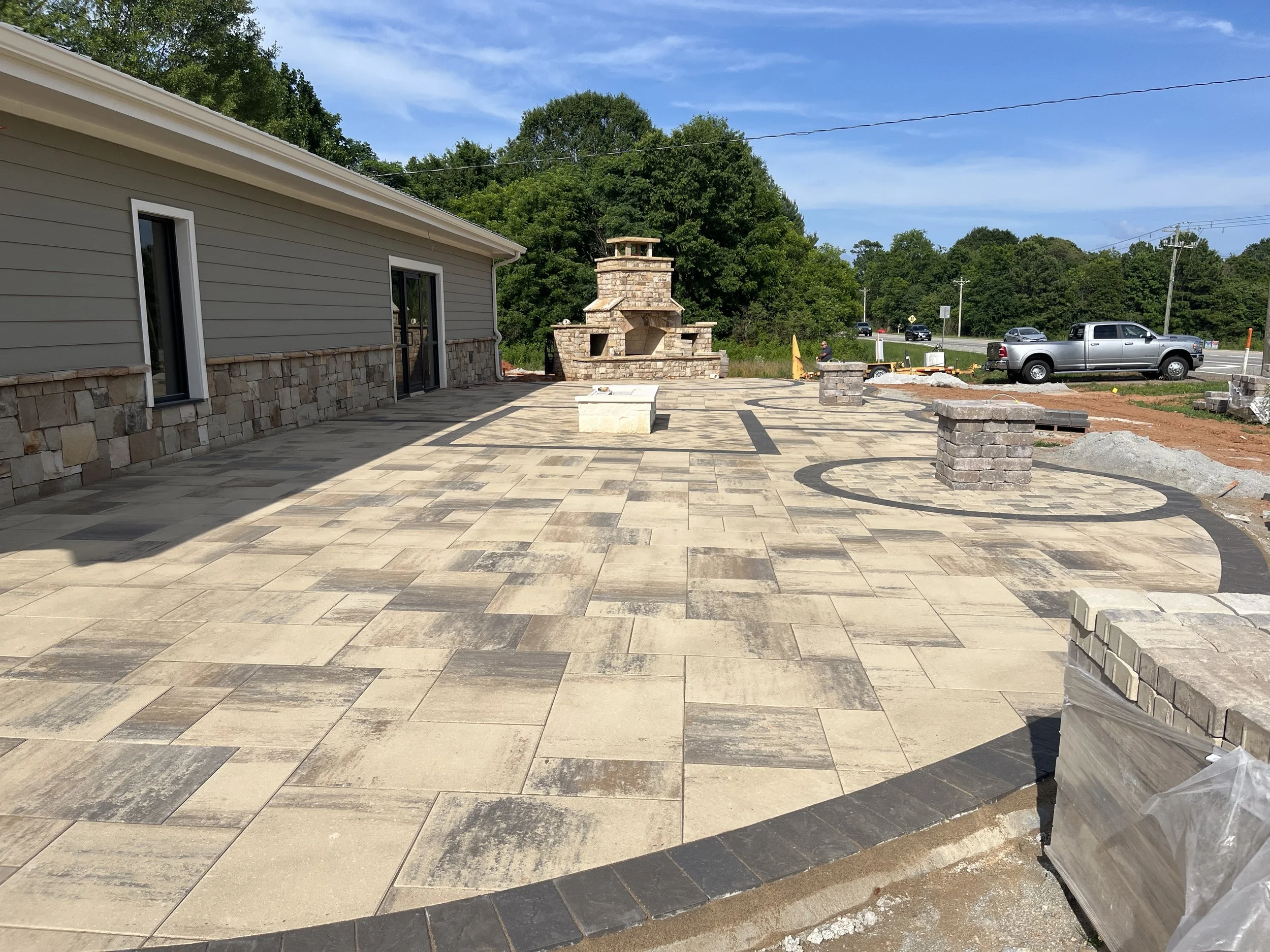 A newly paved outdoor patio area with patterned stone flooring, a brick outdoor fireplace, and a house with beige siding and stone accents. Construction materials and equipment are visible, indicating ongoing work.