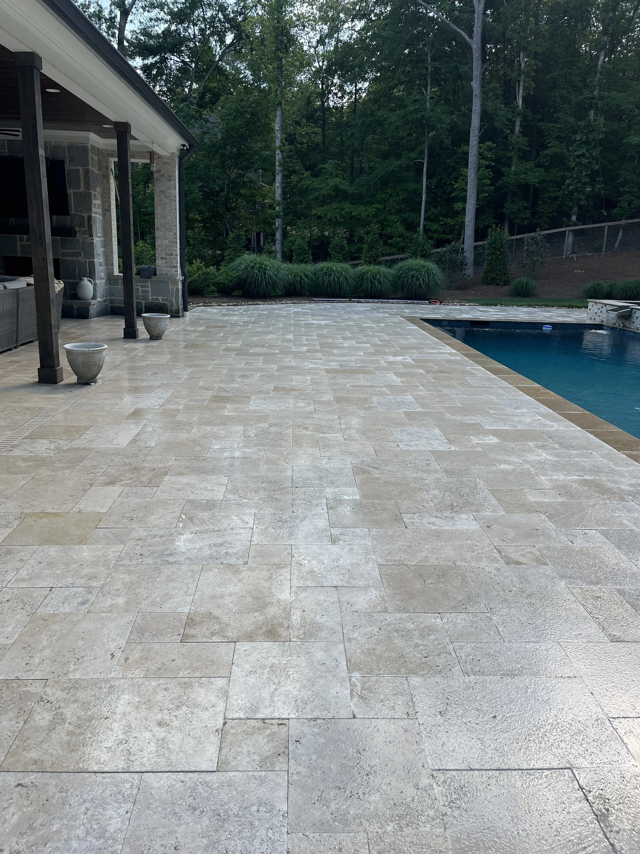 Paved patio area next to a house with a stone facade, white trim, and a ceiling with recessed lights. There are three decorative planters on the patio, and a swimming pool on the right side. In the background, there is a wooded area with tall green t