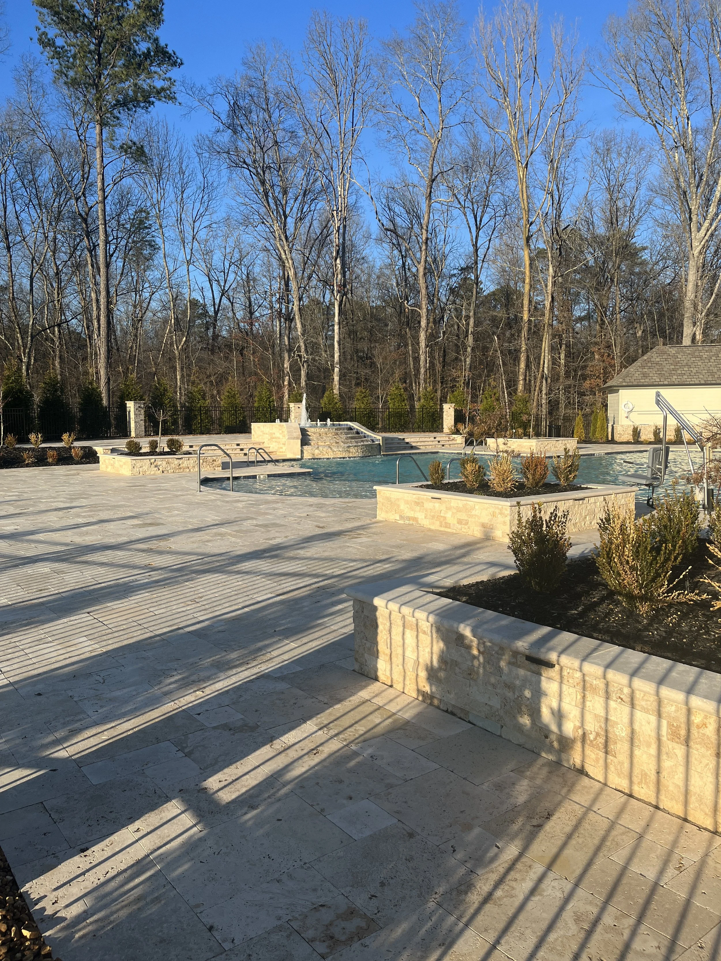 A swimming pool area with stone tile decking and planters with small bushes, set against a backdrop of leafless trees and a bright blue sky.