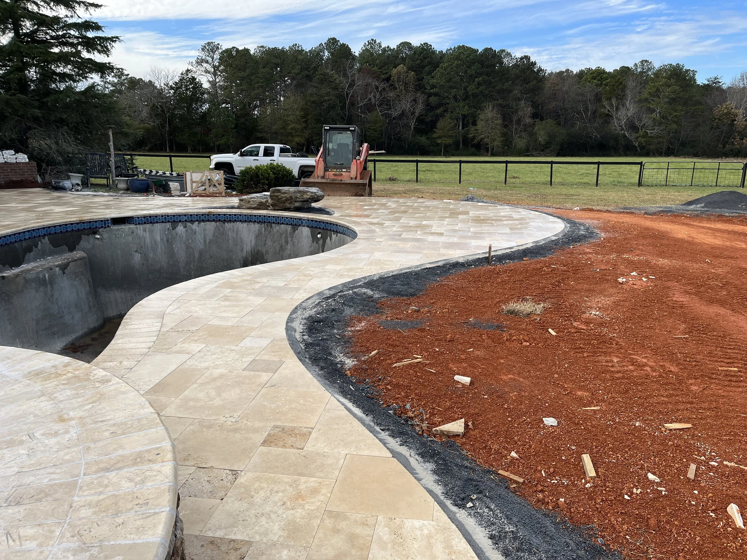 A backyard scene shows a pool under construction with a landscape of trees and a grassy field in the background. Construction equipment and materials are scattered around, and part of a paved area is visible in the foreground. There's a clear sky wit