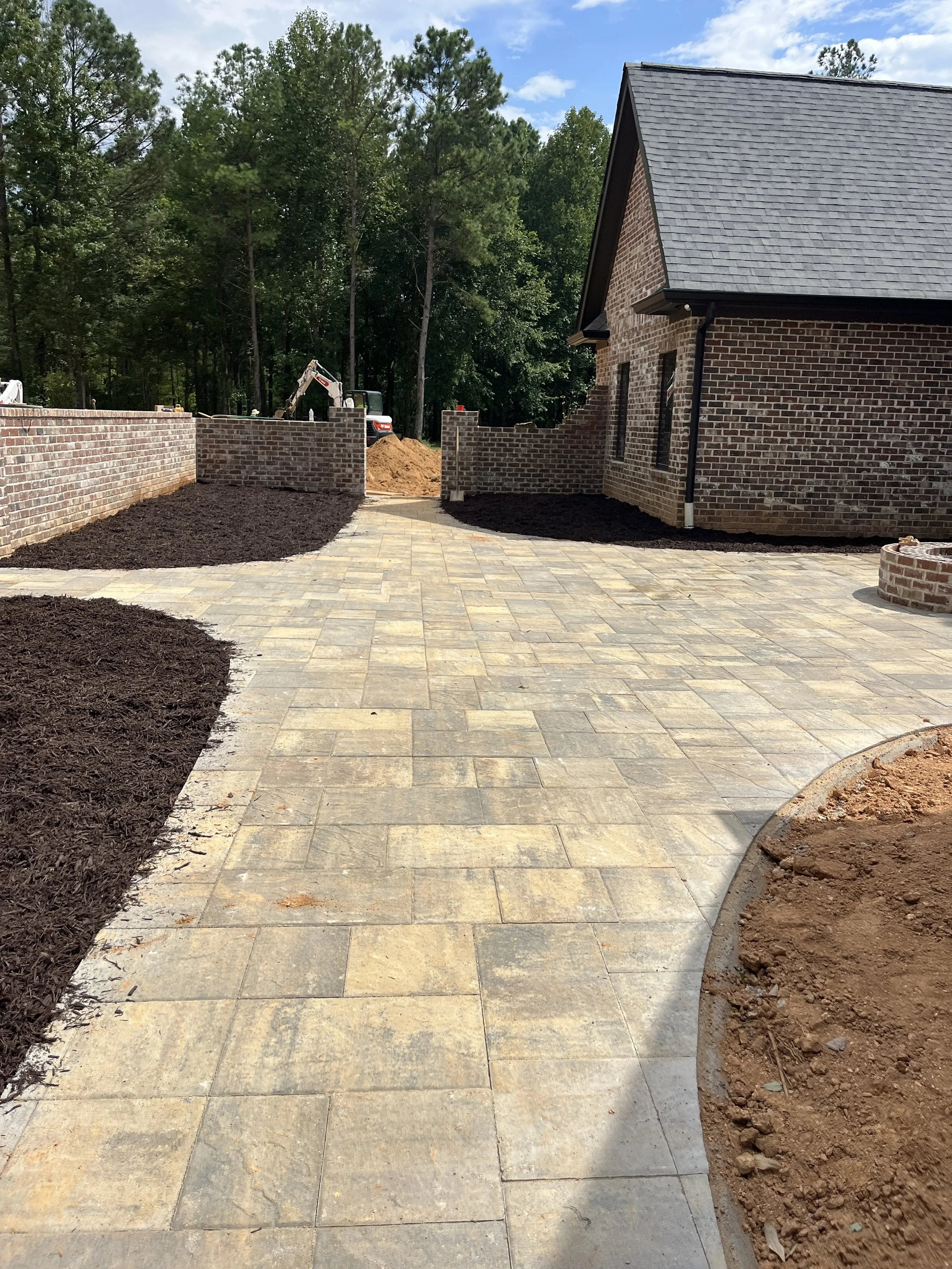A newly paved stone patio area next to a brick house with a black shingle roof, during construction with garden beds and a small excavator in the background, surrounded by trees and a partly cloudy sky.