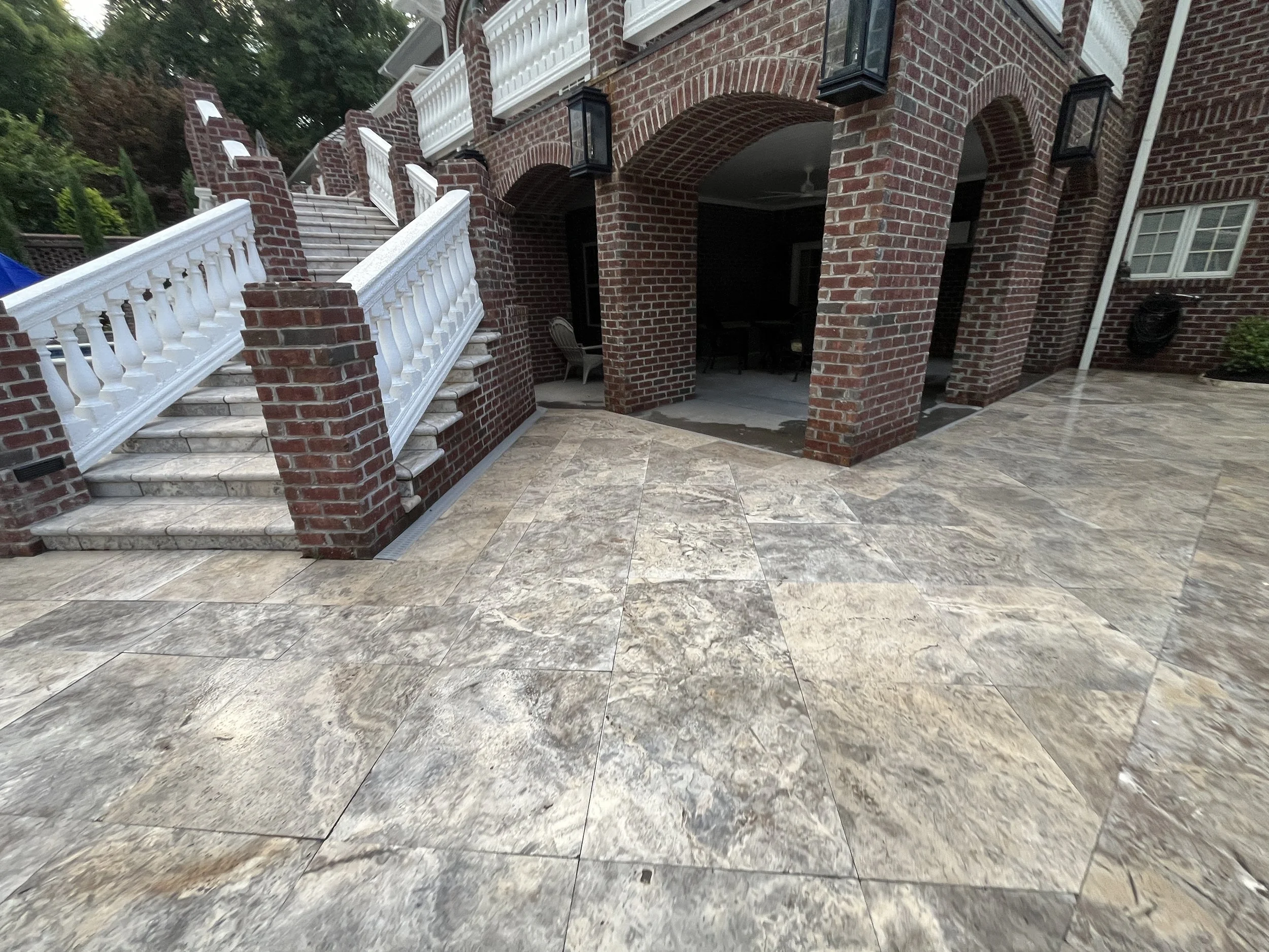 The image shows a brick building with an outdoor patio area. The patio features stone tiles, a staircase with white railings and balusters, brick arches, and black outdoor lighting fixtures.