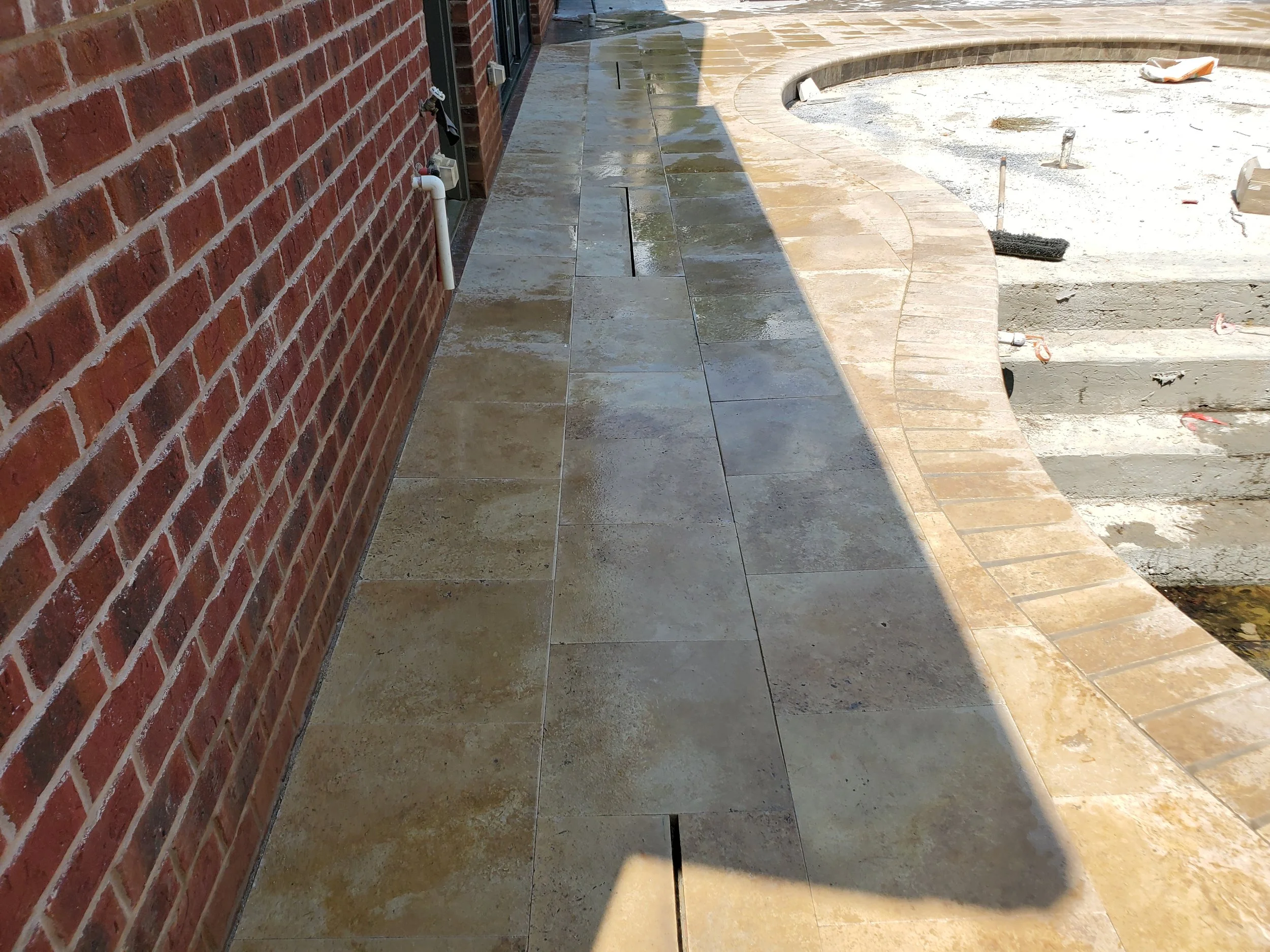 Photo of a partially finished outdoor patio area with new stone pavers and a curved brick wall, construction tools and materials visible.