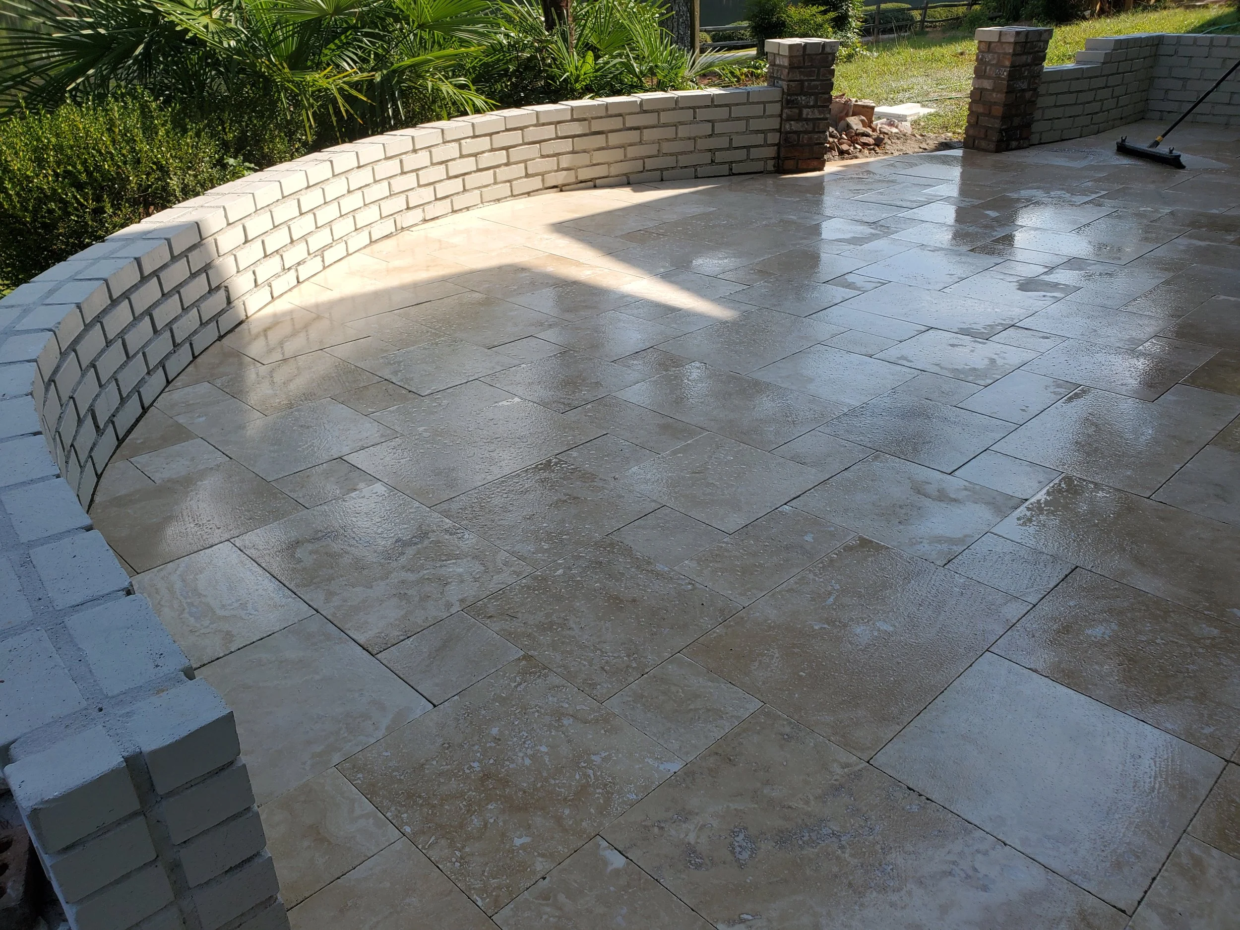A newly constructed outdoor patio with stone tiles and a curved brick wall, partly wet, with a broom leaning against a brick column and greenery in the background.