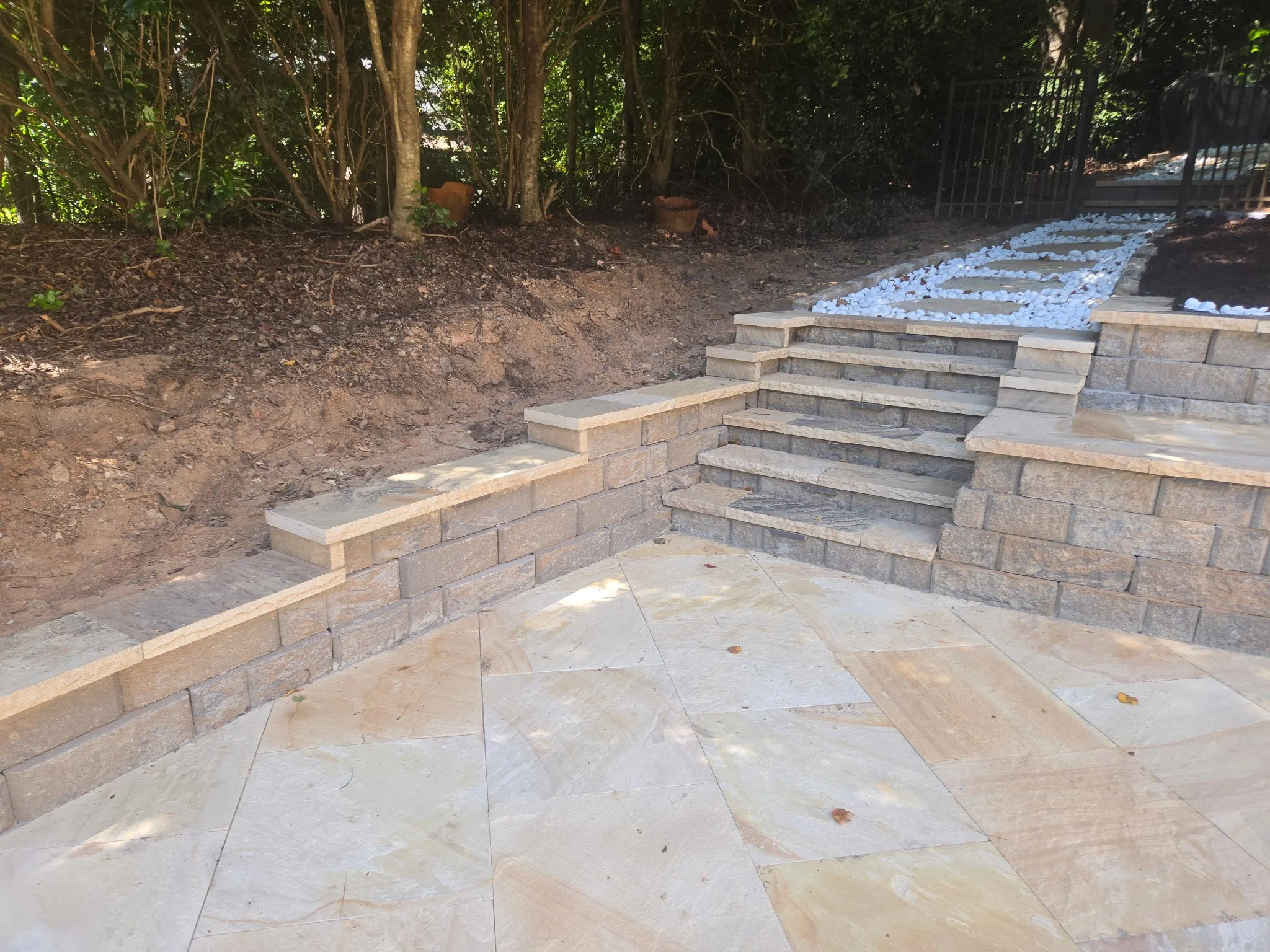 Concrete steps leading up to a gated backyard area with white decorative stones and a wooded background.