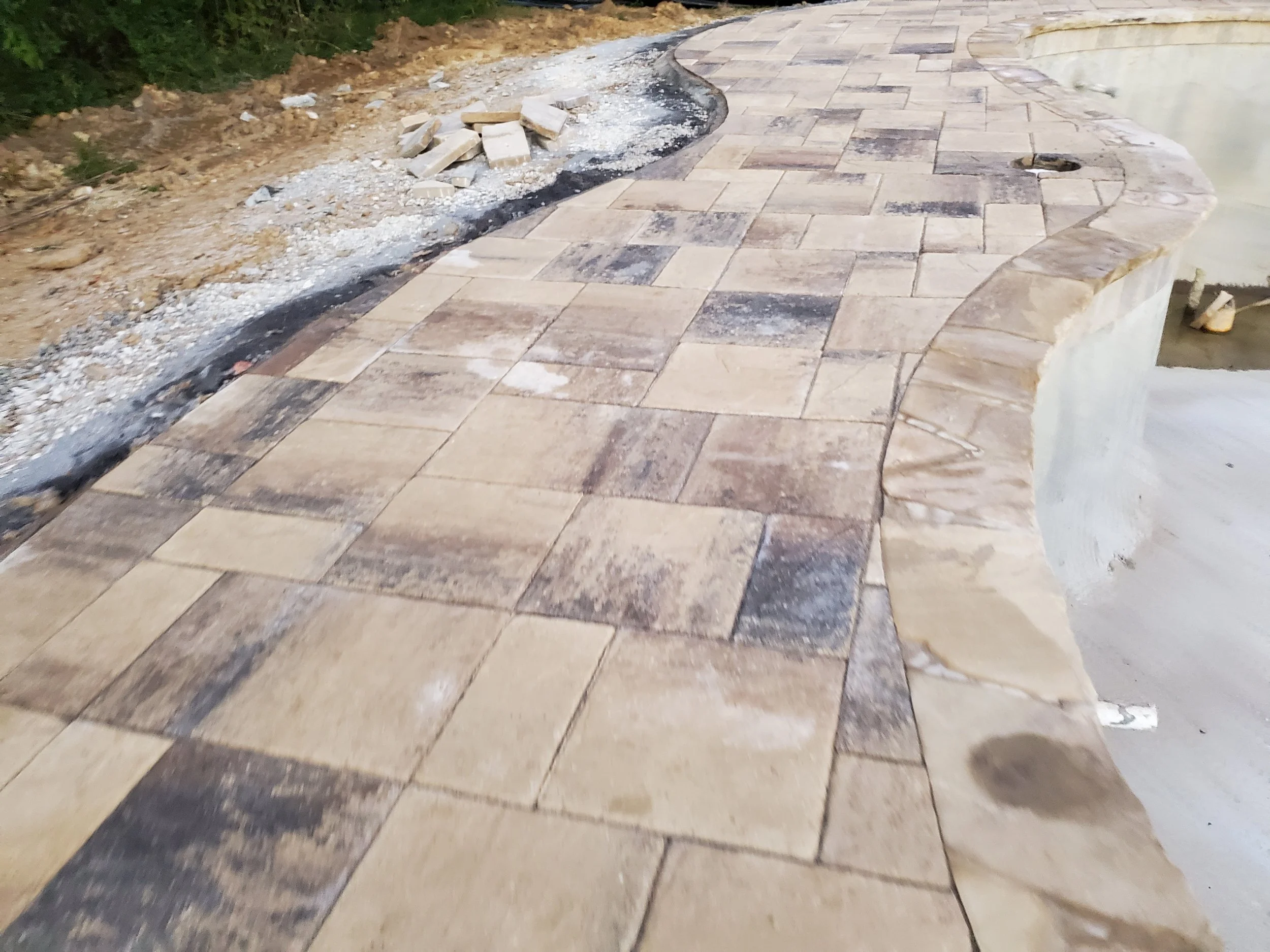 Partially completed outdoor stone paving with scattered bricks and construction debris, next to a grass and dirt area.