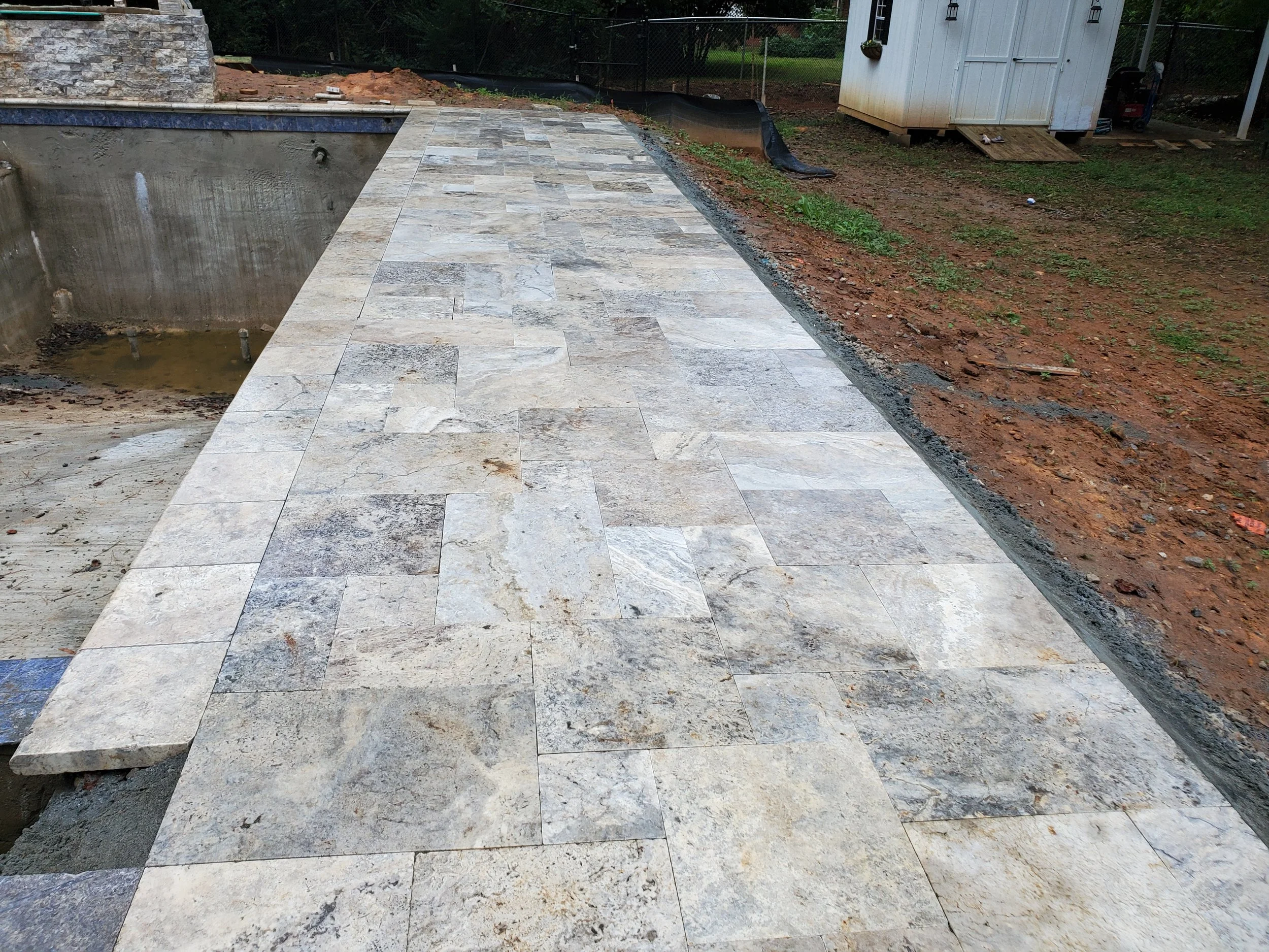 A partially completed outdoor patio with beige and gray rectangular tile pavers, surrounded by exposed soil and construction materials.
