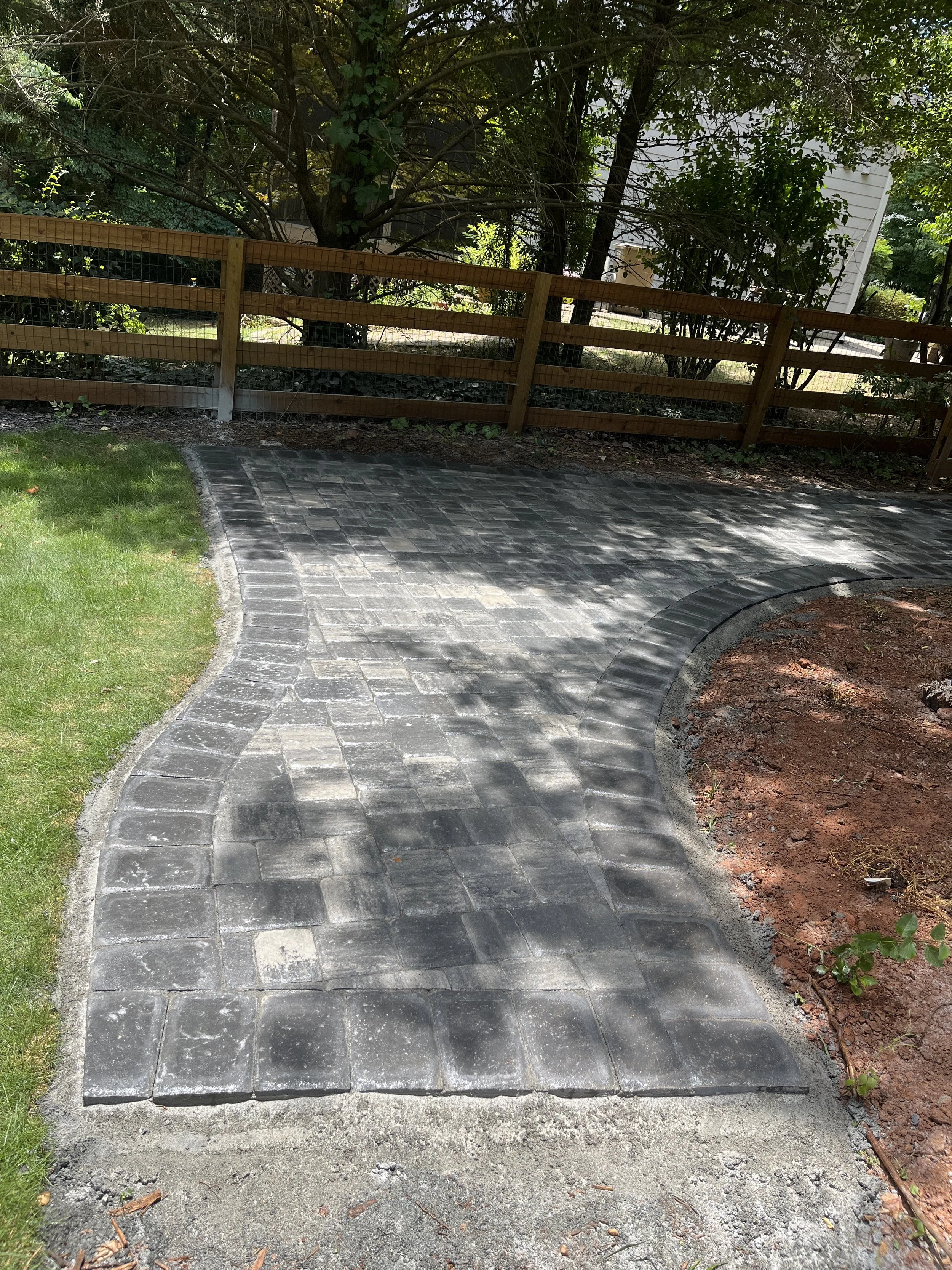 A partially completed black pavers pathway with a curved design, bordered by grass on the left and dirt on the right, with a wooden fence behind shrubbery and trees.