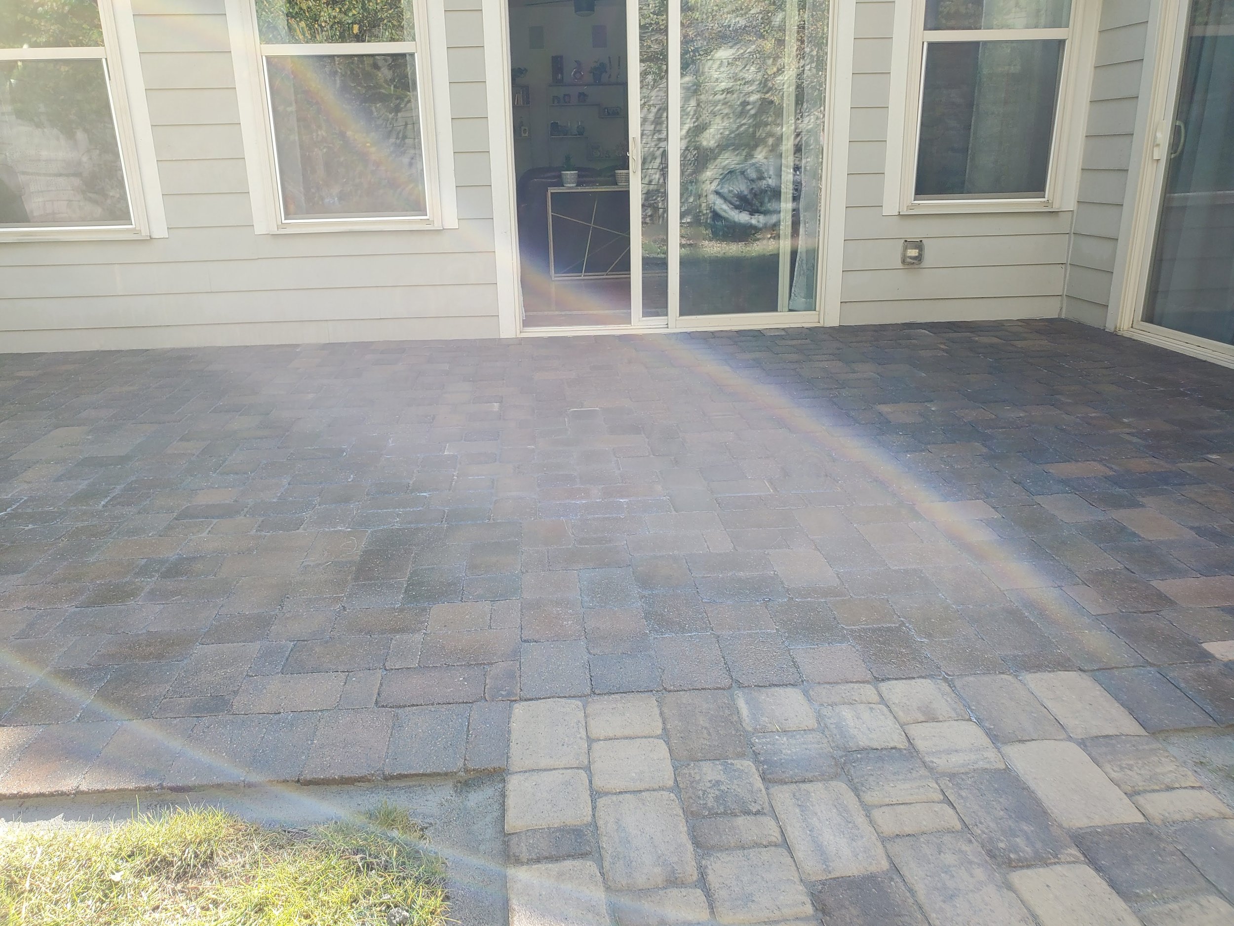 Backyard patio with stone pavers and a glass sliding door leading into a house, seen through the door are bookshelves and furniture inside the house. Sunlight and a rainbow lens flare are visible.