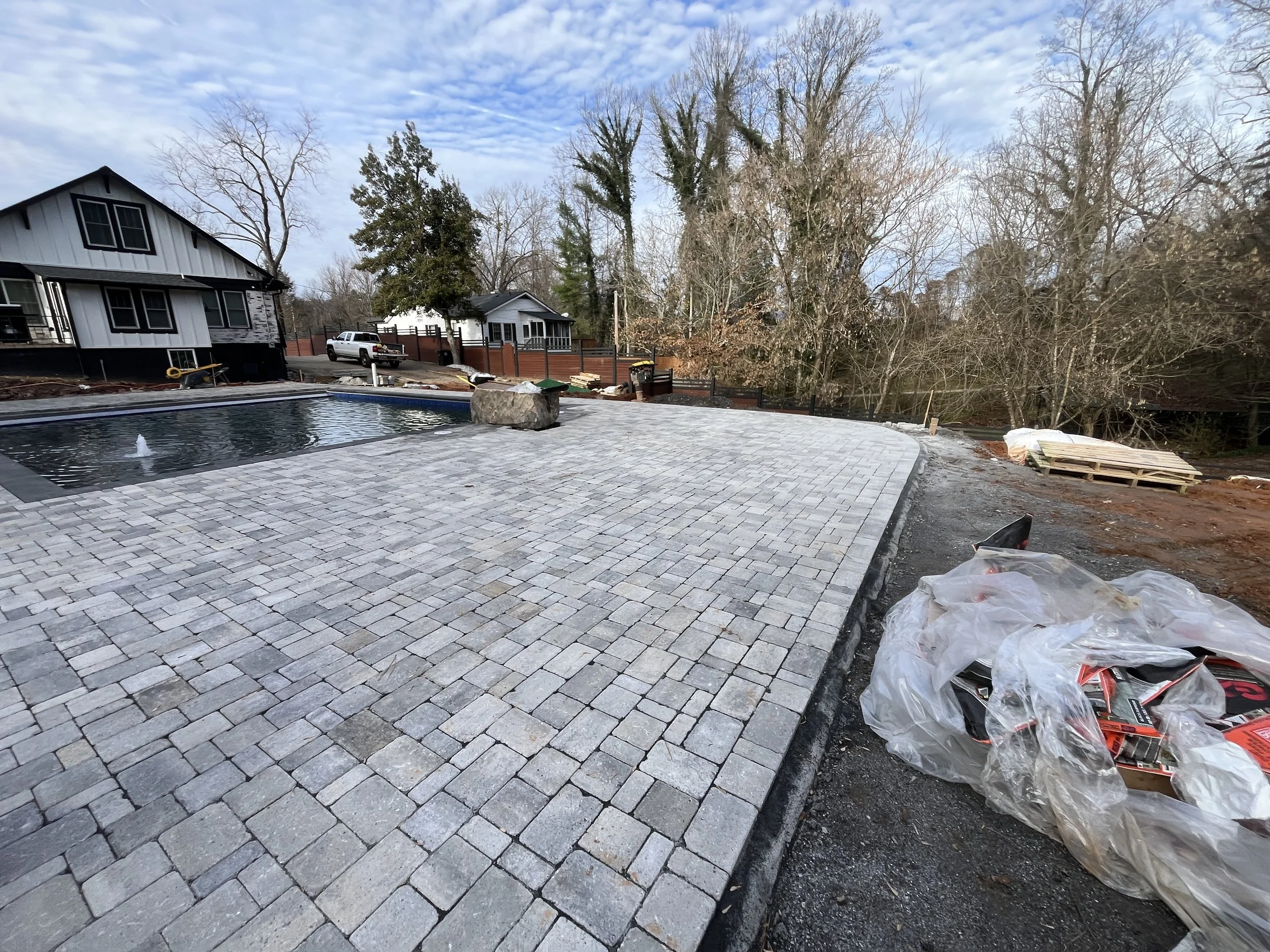 A backyard with a newly paved stone patio, a small water fountain, and a fire pit. There's a white house with black trim and a pickup truck parked near a wooden fence. Bare trees and a cloudy sky are in the background.