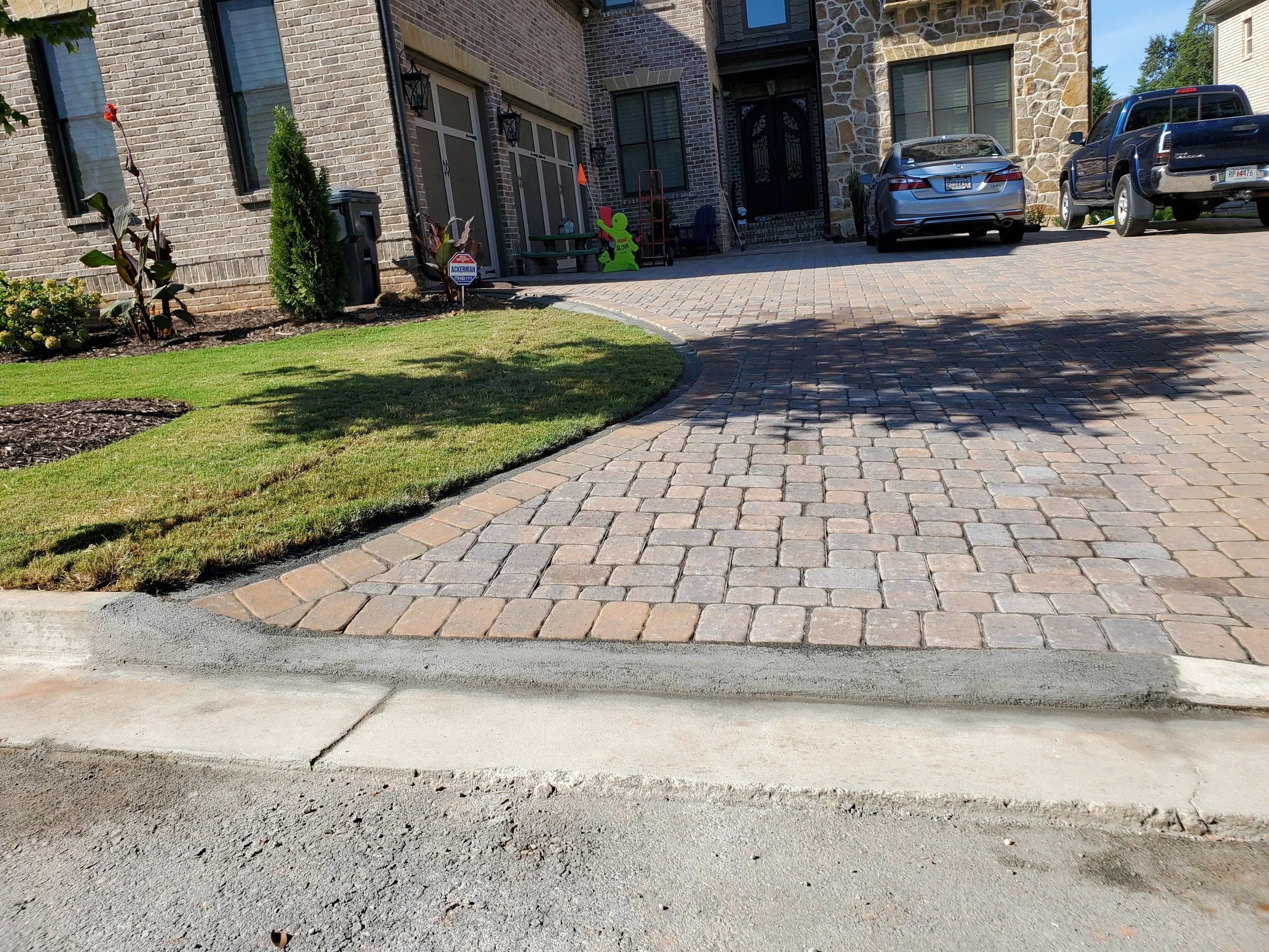 The image shows a paved driveway in front of a house with plants and a lawn on the left and cars parked on the right side. The house has brick walls, windows, and a black front door, with various outdoor items near the entrance.