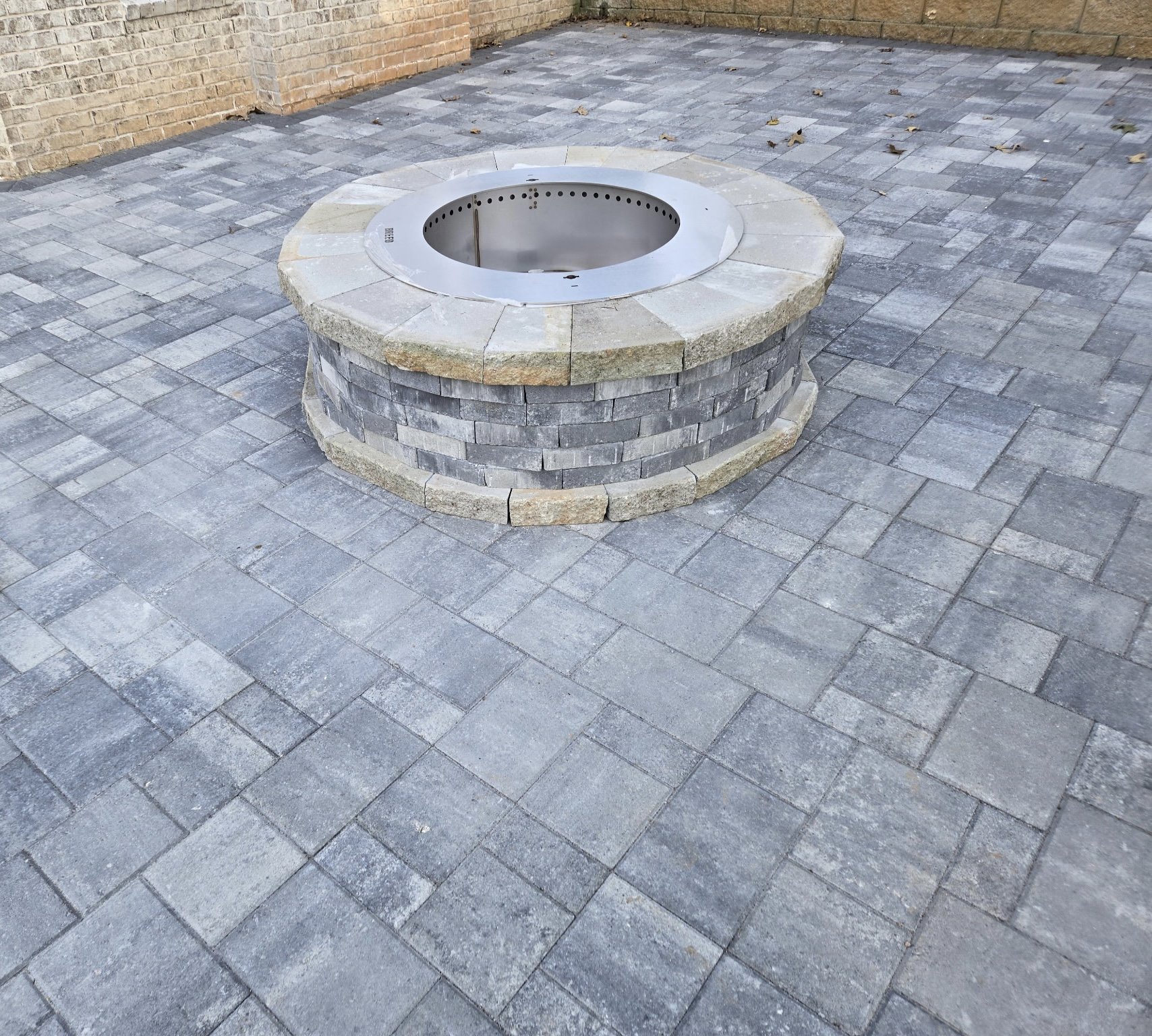A fire pit with a round brick base and a metal insert, surrounded by gray brick pavers on an outdoor patio.