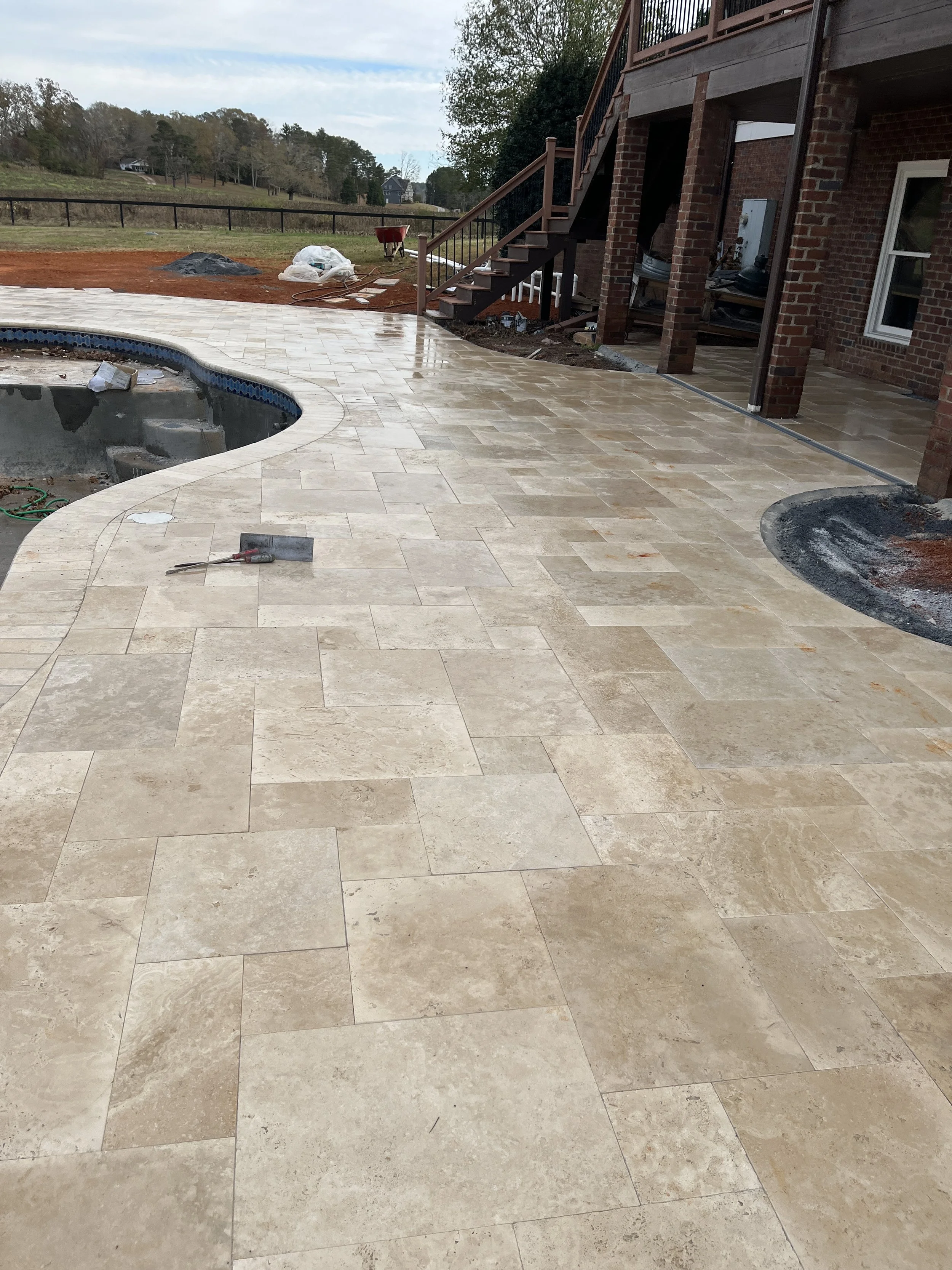 Backyard patio area with beige stone tiles, a swimming pool under construction, and an incomplete brick house construction with tools and materials scattered around.