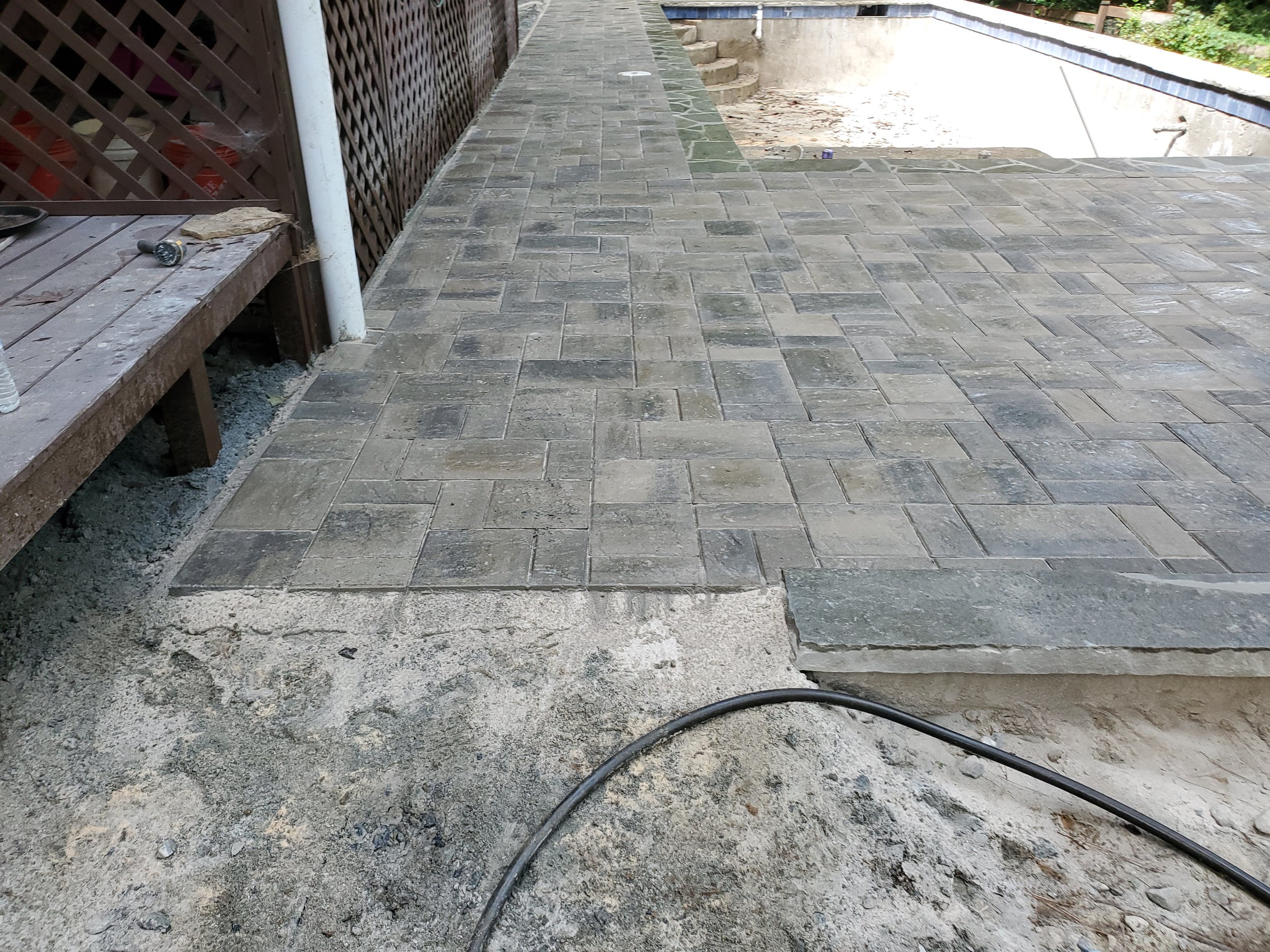 A partially completed brick patio or walkway with some areas still under construction, including exposed gravel and sand base, a black hose laying on the ground, and a wooden bench with tools and debris on it to the left, alongside a wooden lattice f