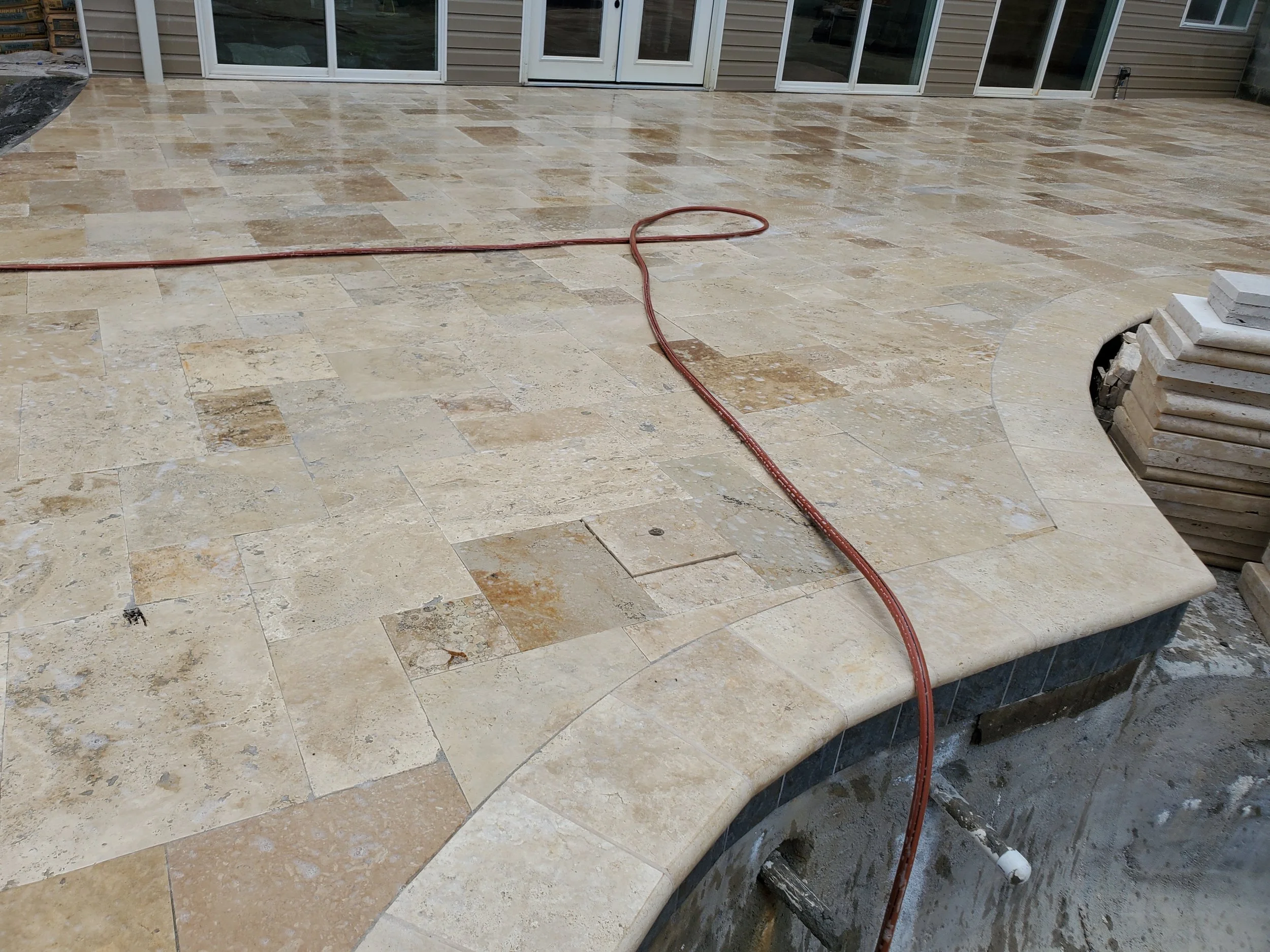 A newly installed light-colored stone or ceramic tile patio outside a house with sliding glass doors and a single door. There are construction materials and a red extension cord on the patio.