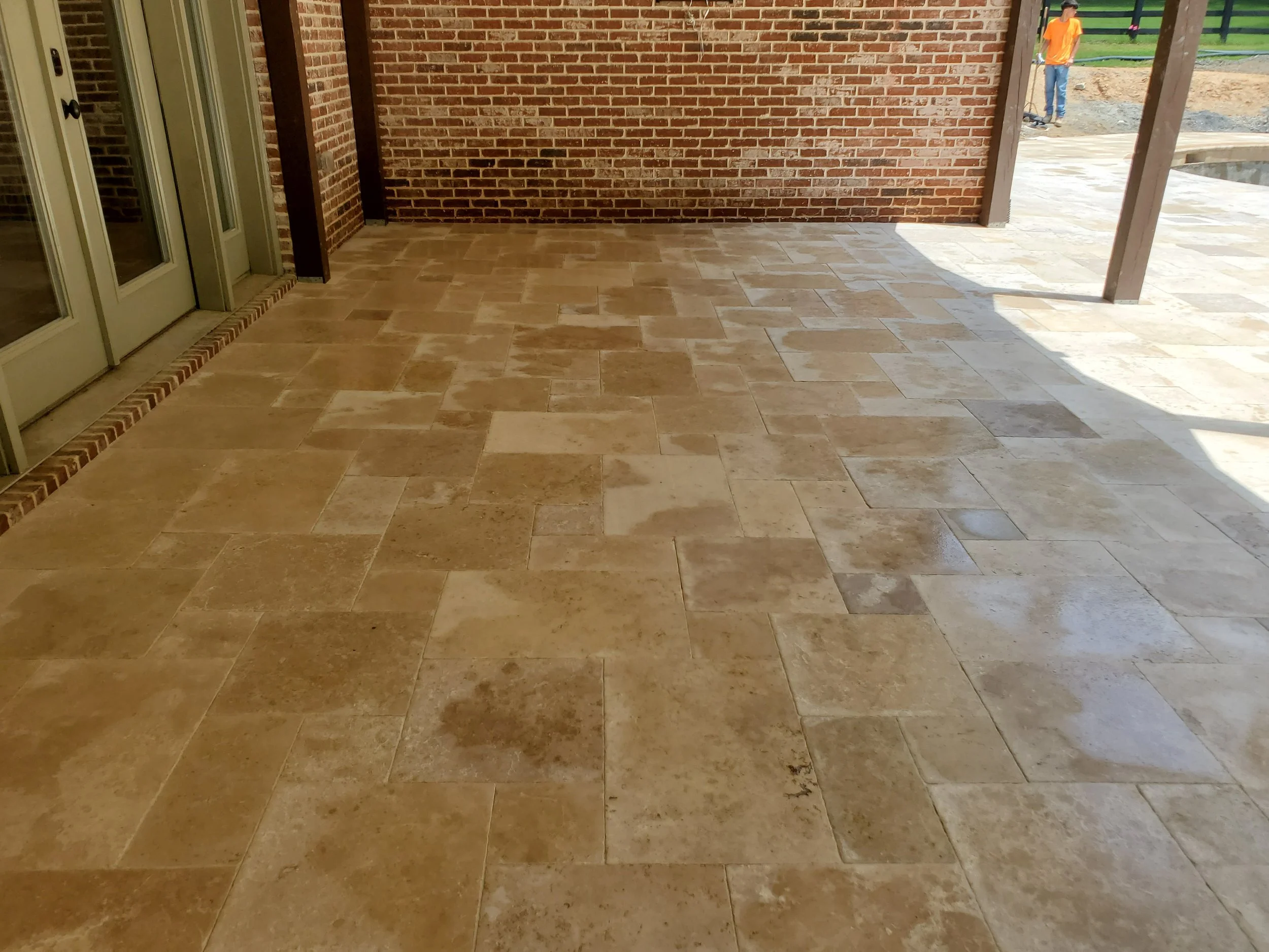 An outdoor patio with cream-colored stone tiles, brick walls, and a sliding glass door leading inside. Two wooden support beams are visible on the right, with ongoing construction visible outside where workers are present.