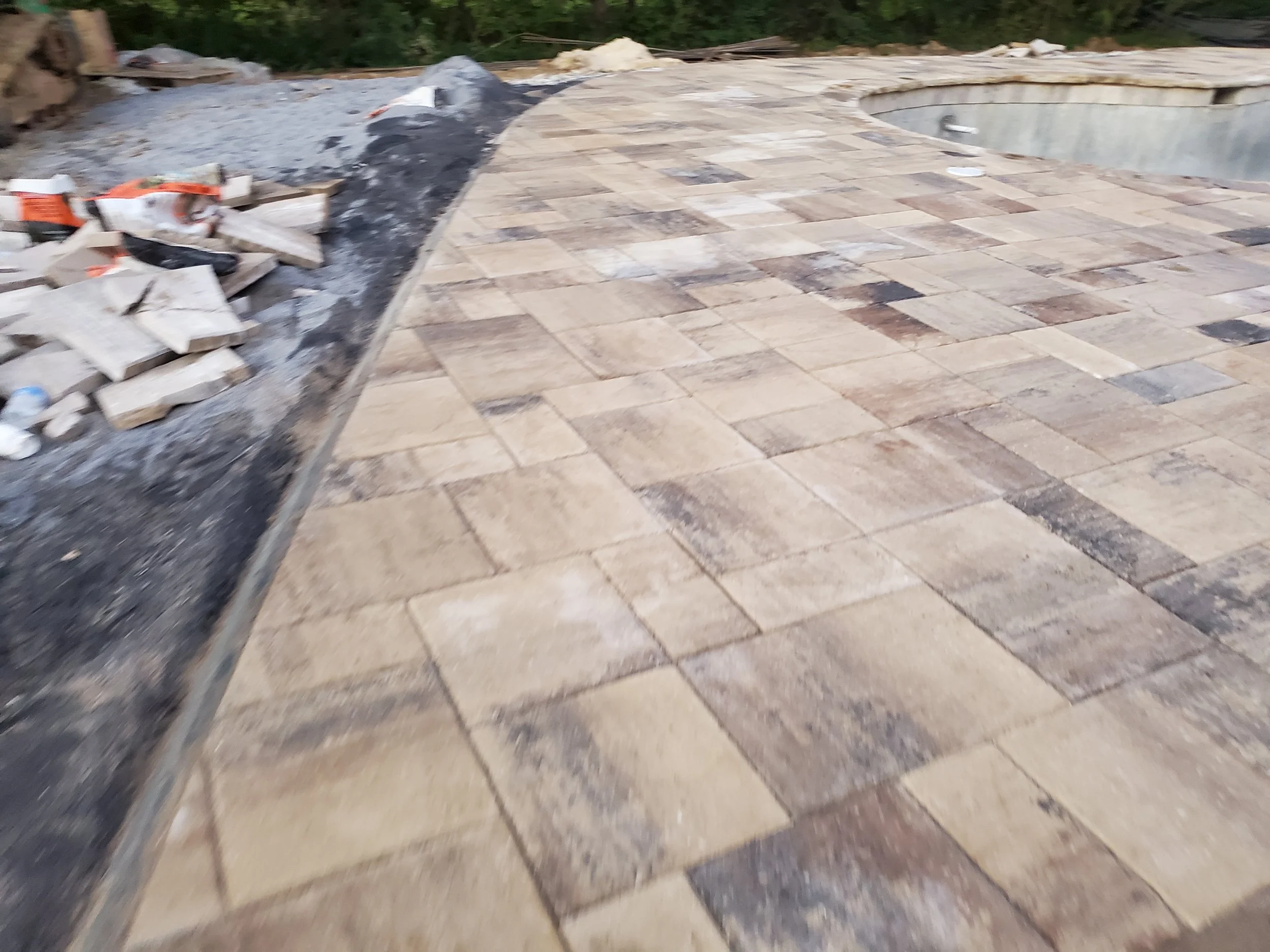 A partially completed outdoor patio with new paving stones and a swimming pool in the background. Construction debris is piled on the side.